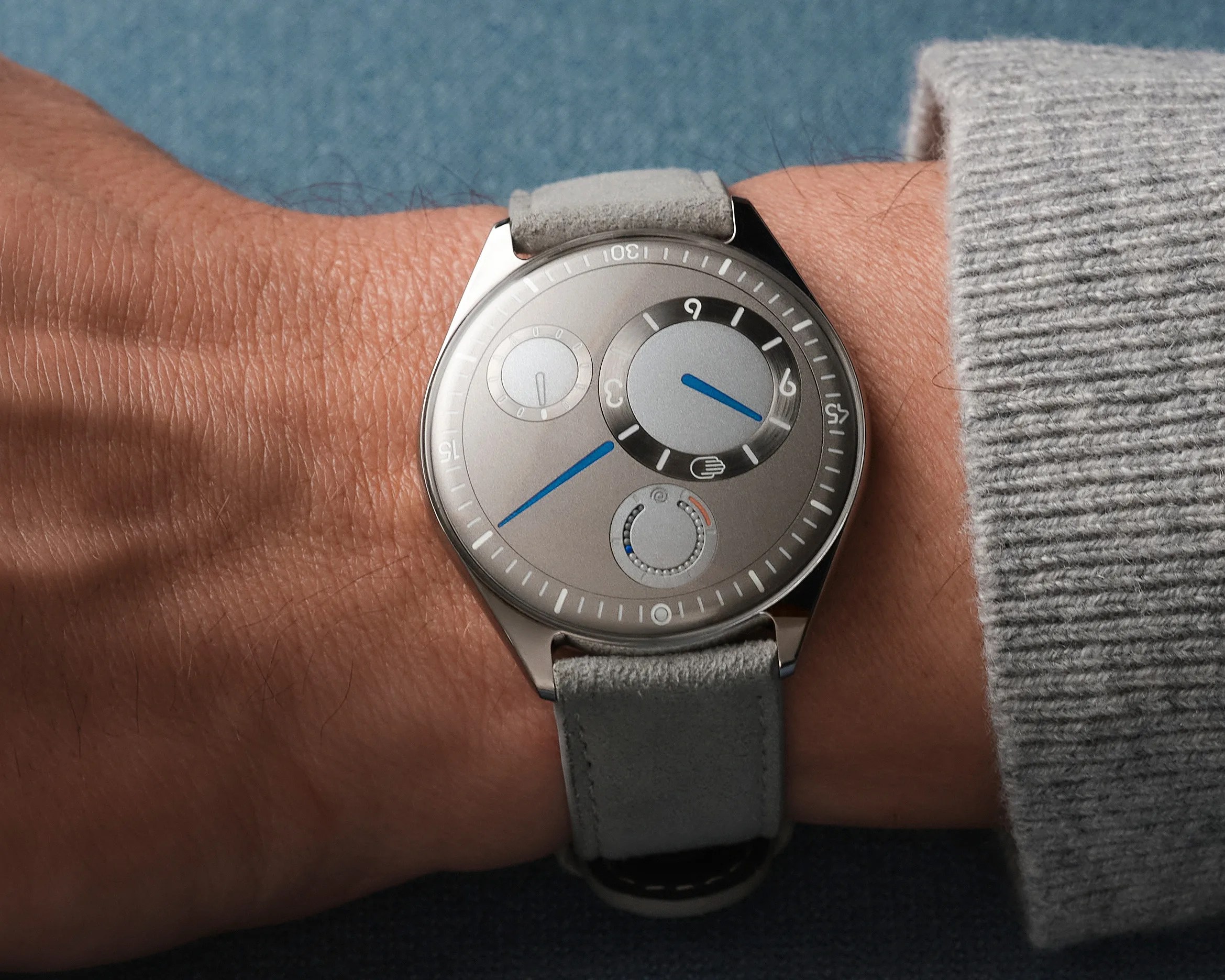 Silver wristwatch with blue hands and gray suede strap worn on a wrist with a gray sweater cuff.