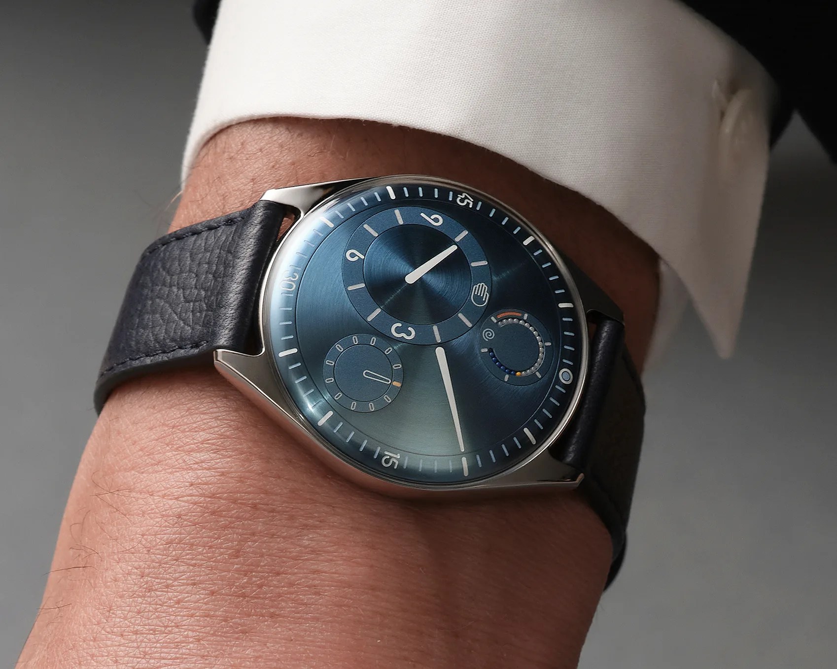 Wristwatch with a blue sunburst dial, silver case, and black textured leather strap worn on a wrist.
