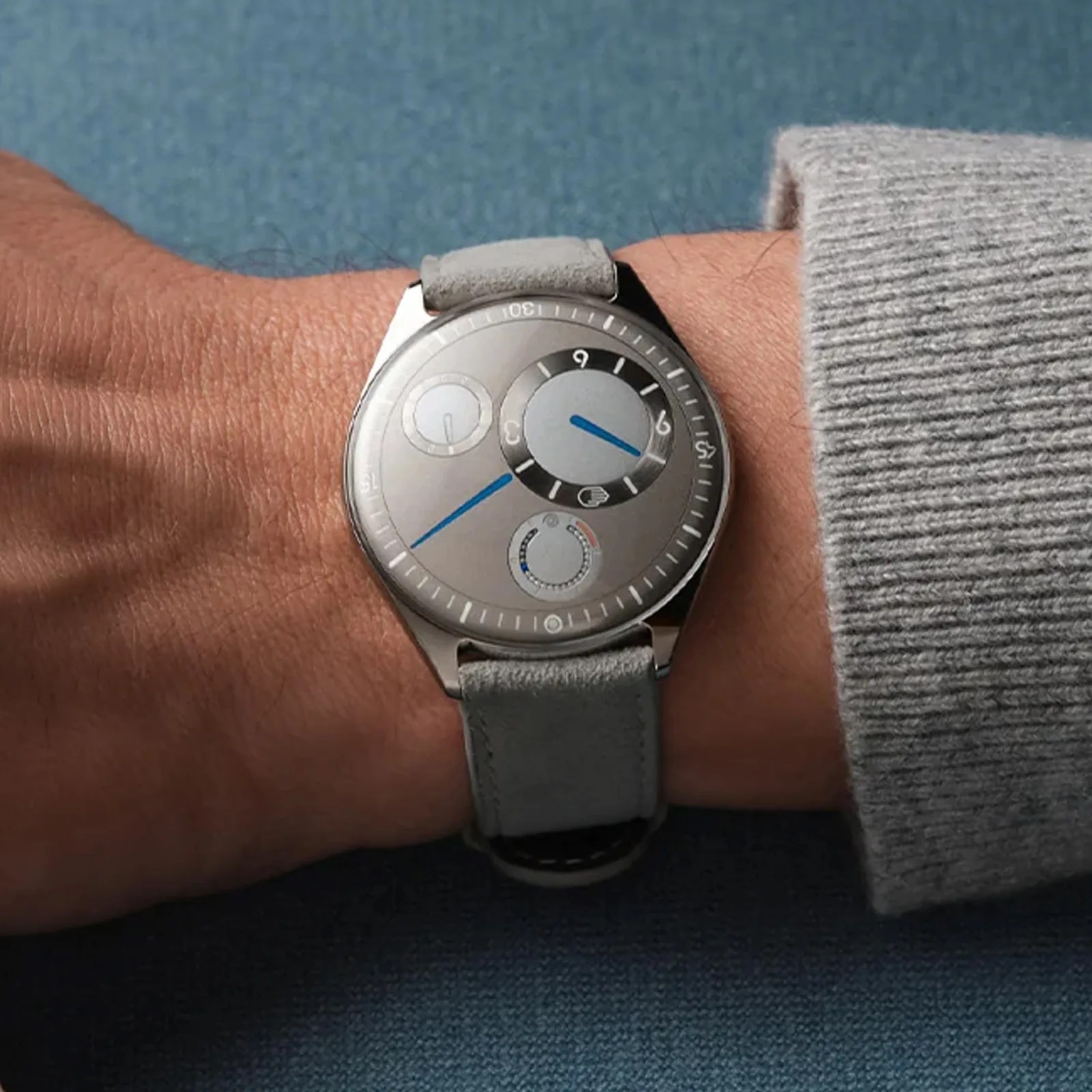 Silver wristwatch with blue hands and gray suede strap worn on a wrist with a gray sweater cuff.