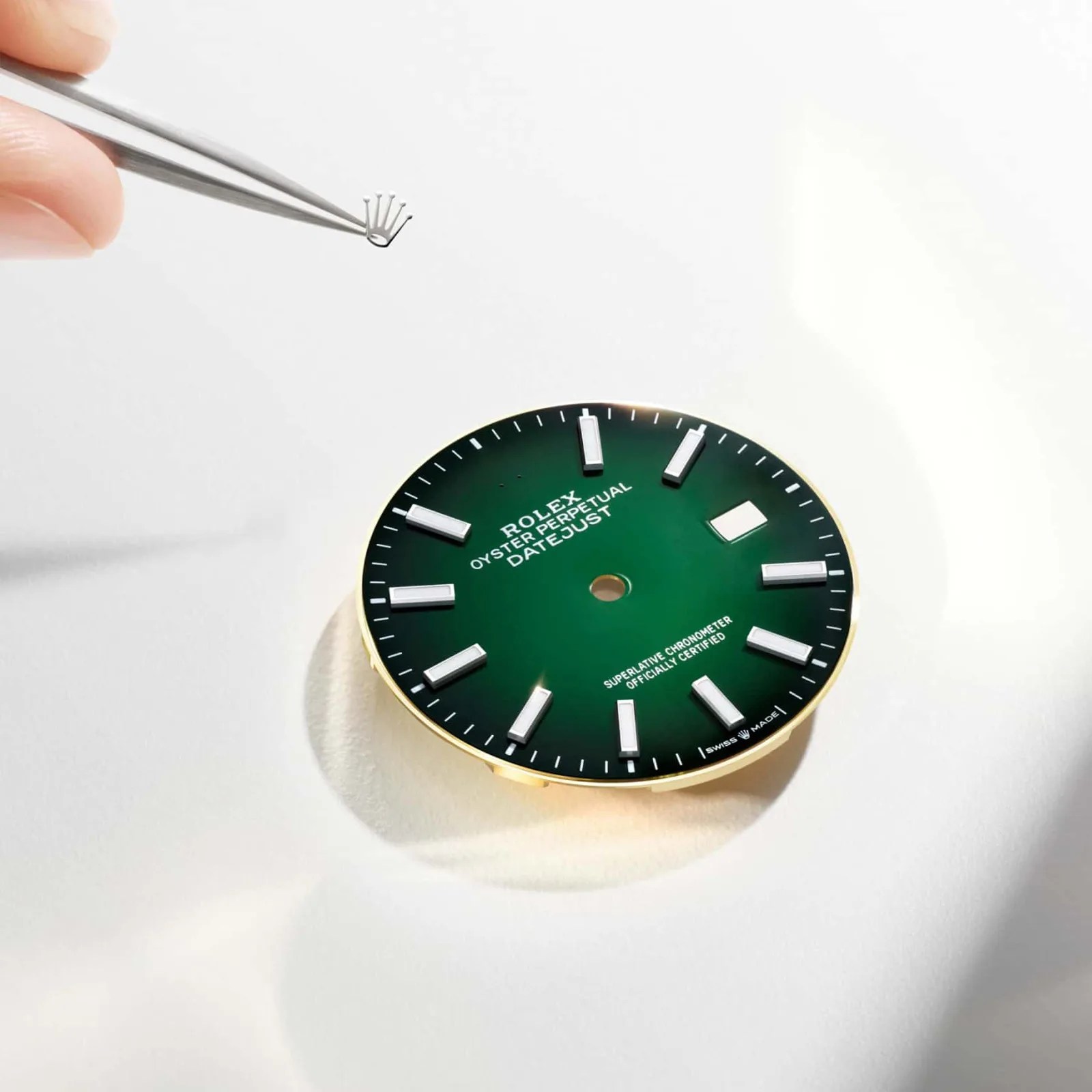 Green Rolex Oyster Perpetual Datejust watch face with silver hour markers and a hand holding a small Rolex crown logo with tweezers.