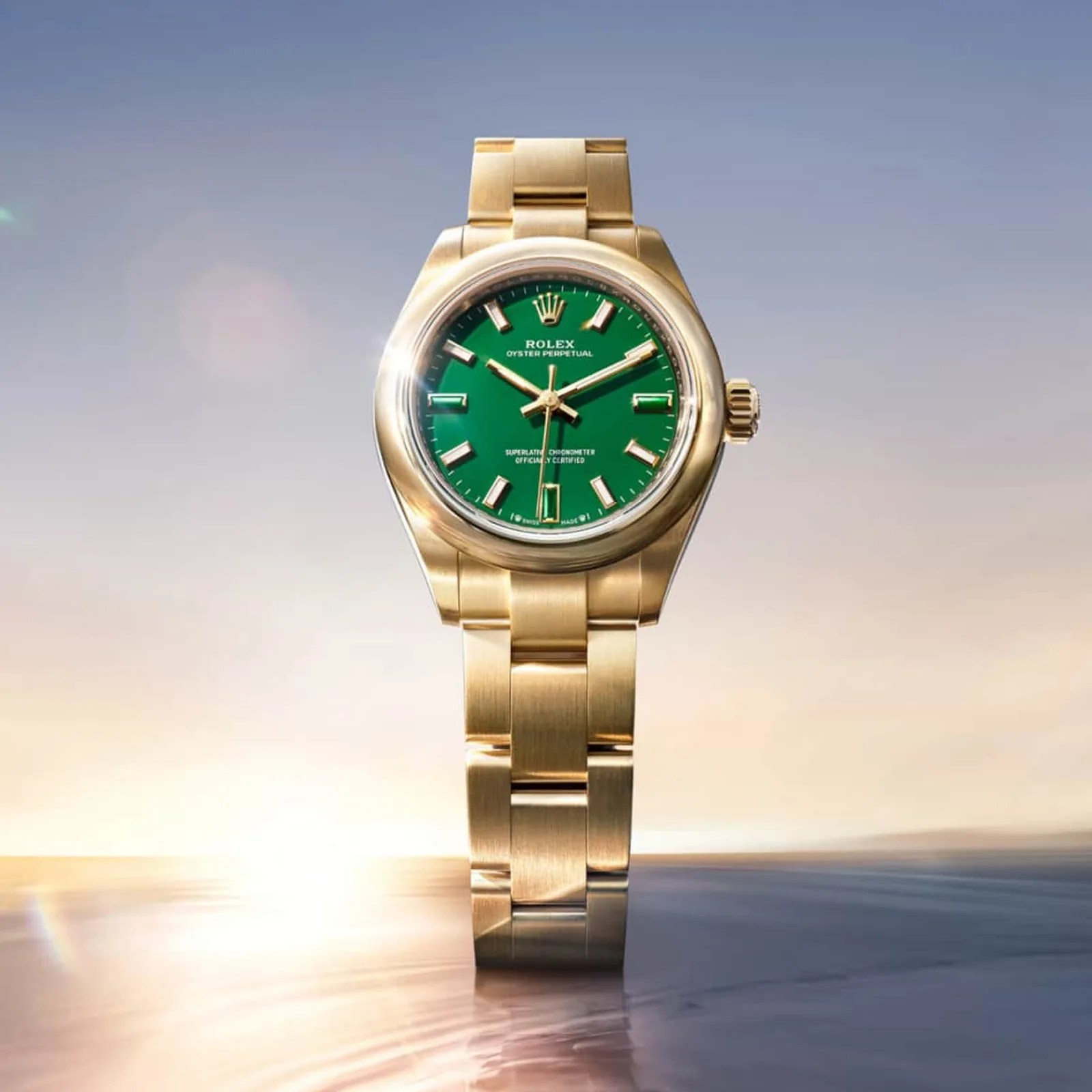 Gold Rolex Oyster Perpetual watch with a green dial and gold bracelet against a blurred sunset background.