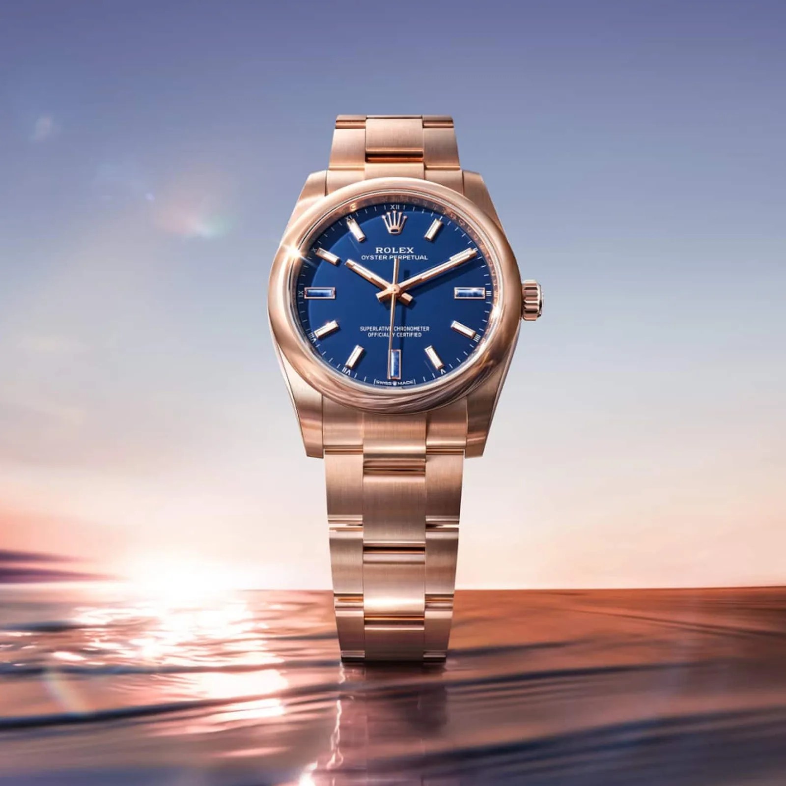 Rose gold Rolex Oyster Perpetual watch with a blue dial standing on reflective water at sunset.