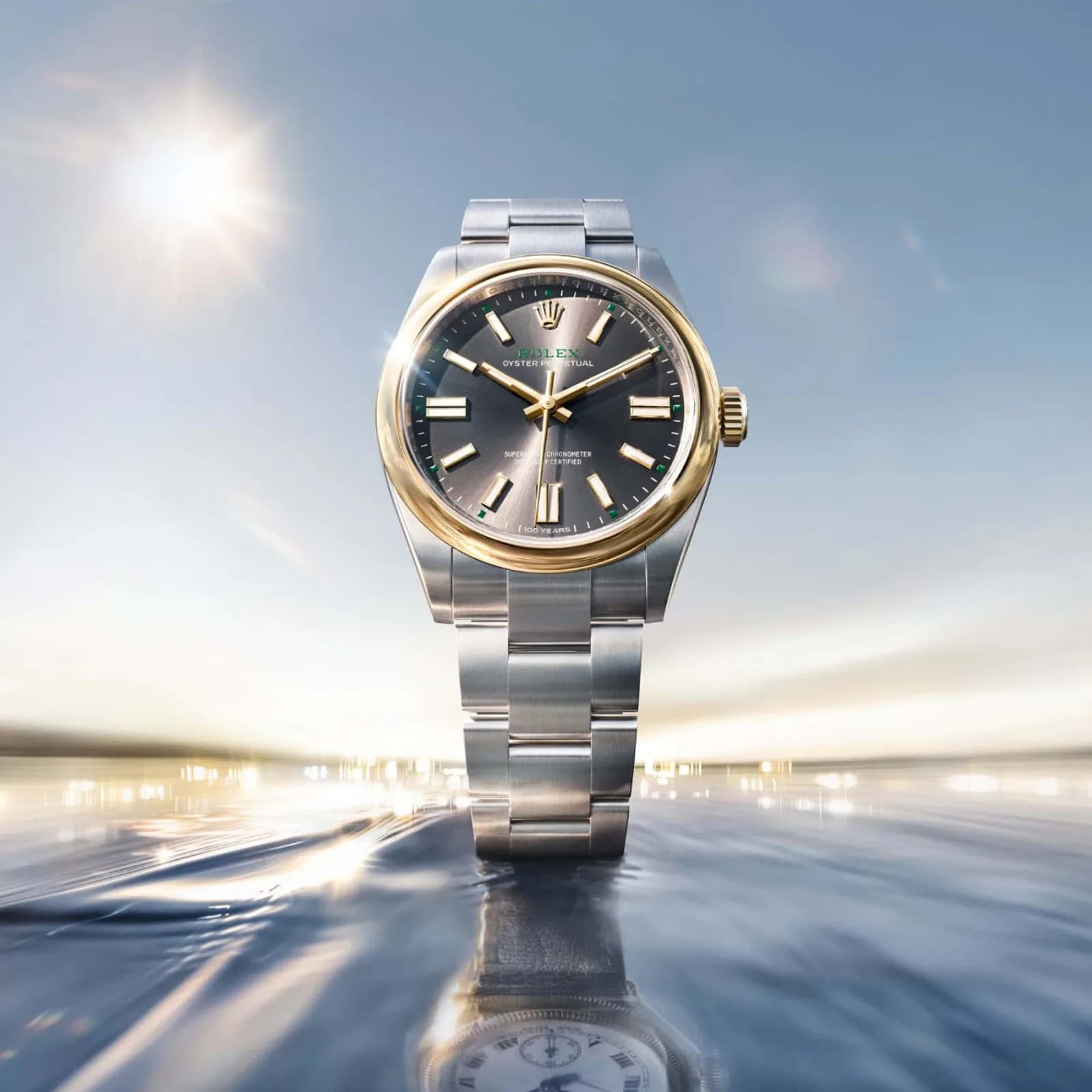 Two-tone Rolex Oyster Perpetual watch with a black dial and gold bezel standing upright on a reflective surface with a bright sky background.