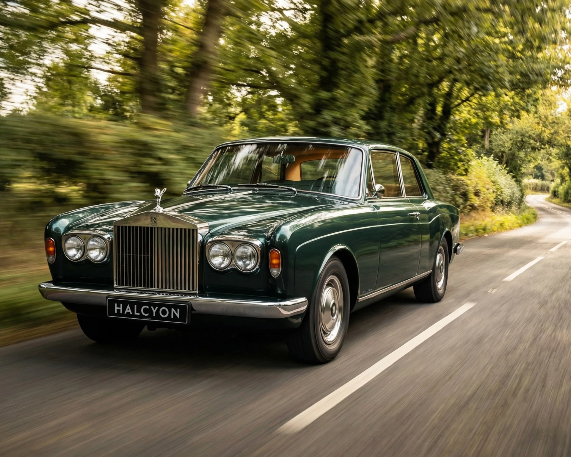 Dark green classic Rolls-Royce car with chrome grille and "HALCYON" plate driving on a tree-lined road.