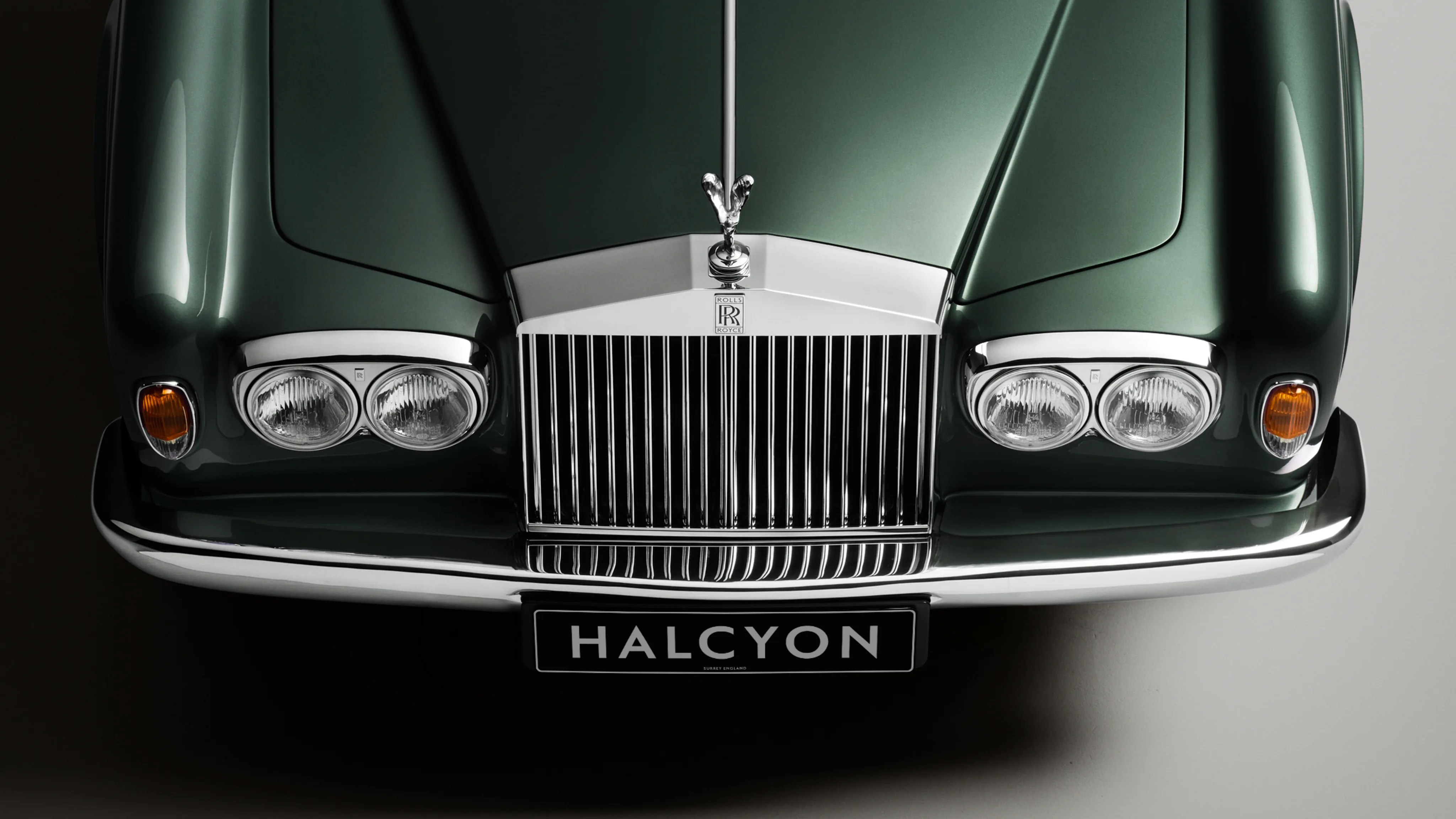 Front view of a dark green Rolls-Royce car with chrome grille, dual headlights, and "HALCYON" license plate.