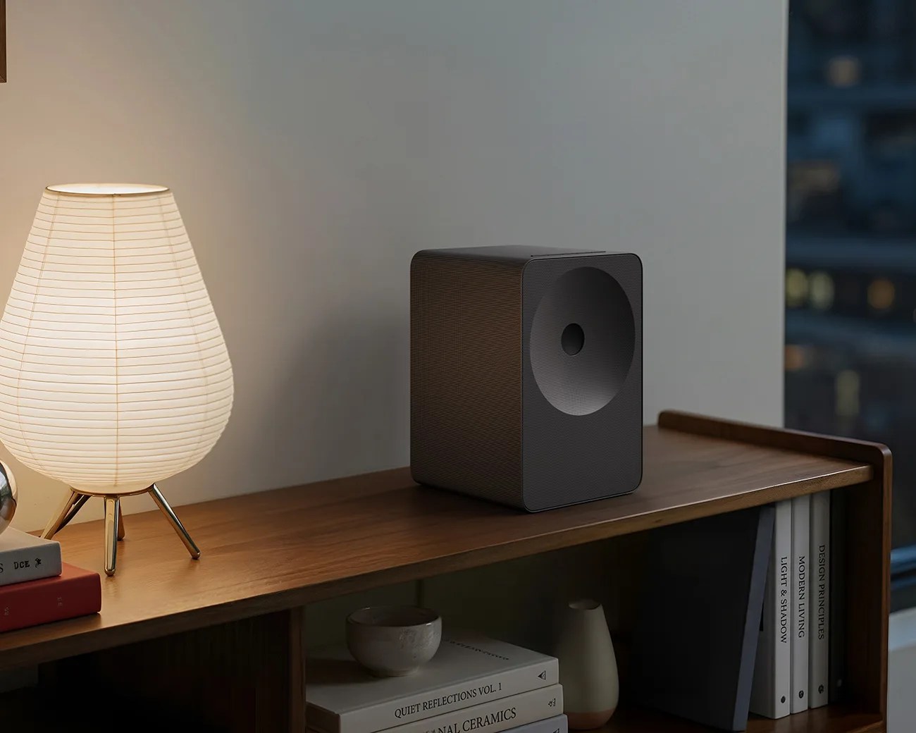 Black rectangular speaker with a circular cone on a wooden shelf next to a lit white paper lamp.