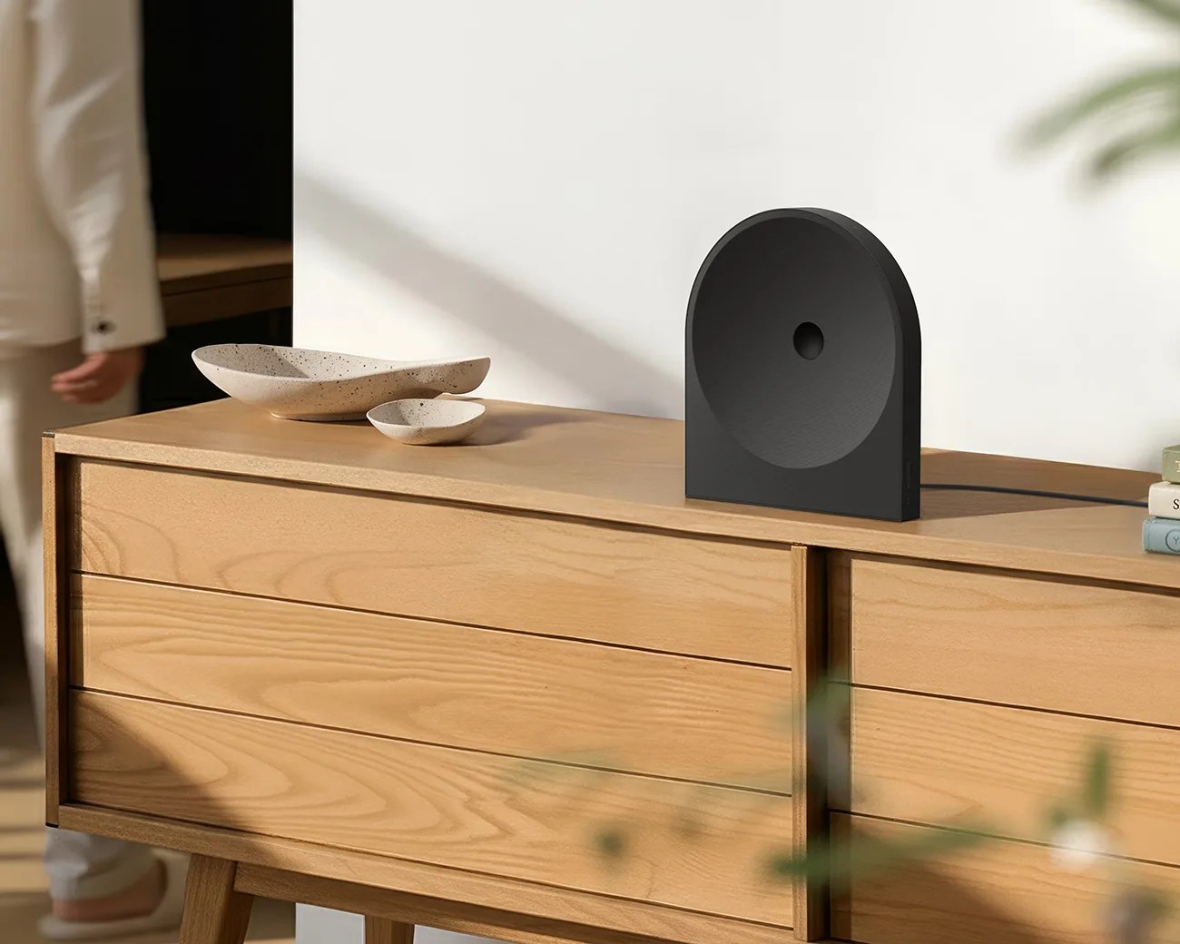 Black semi-circular speaker with a central hole on a light wood sideboard next to two beige ceramic dishes.