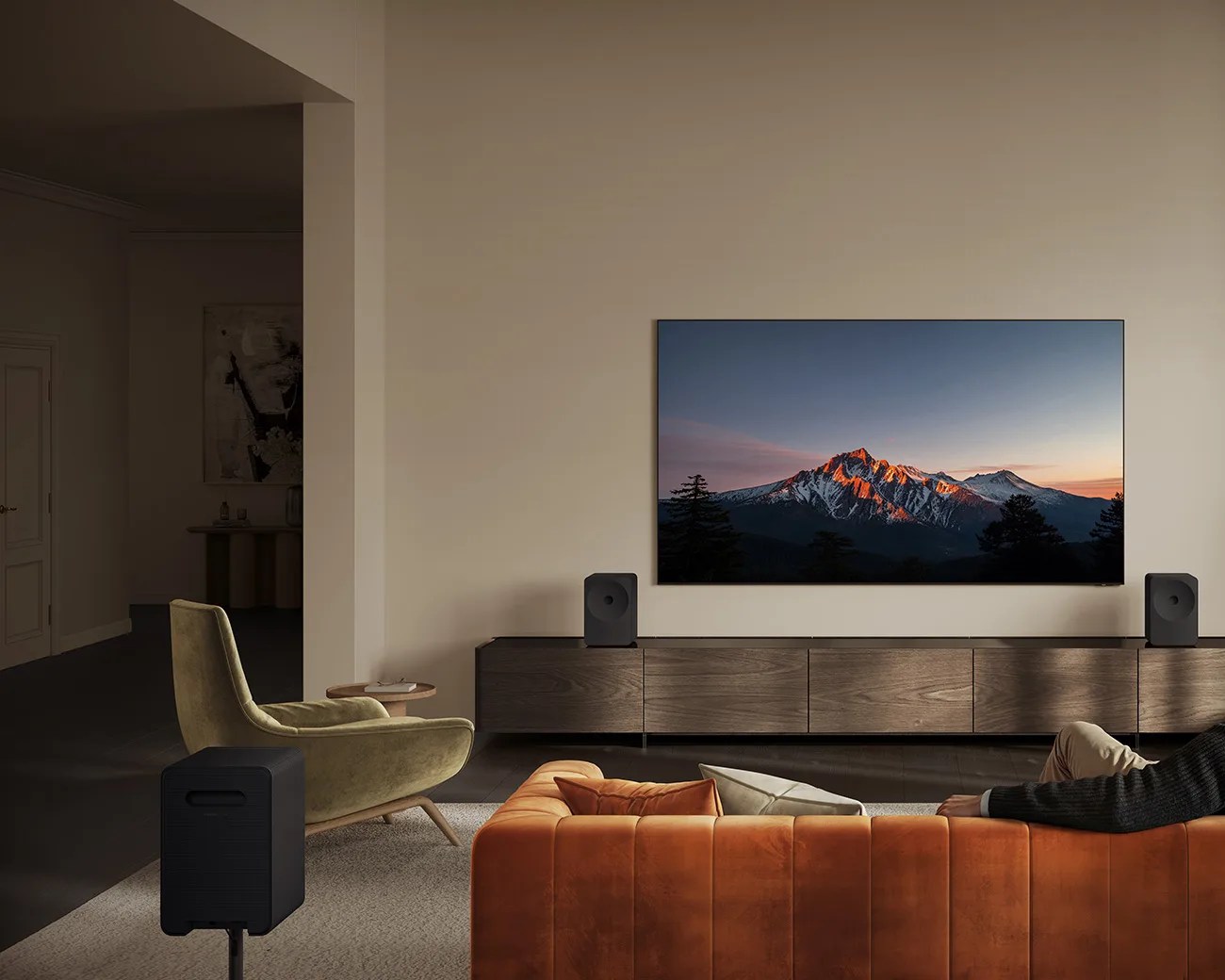 Modern living room with a wall-mounted flat-screen TV showing a mountain scene, two black speakers, an orange sofa, and a green armchair.