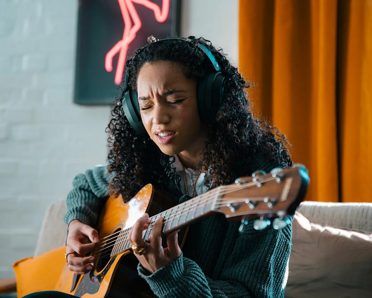 Person wearing black headphones and a green sweater playing an acoustic guitar indoors.