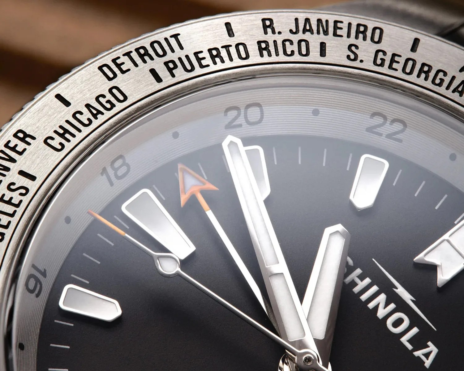 Close-up of a Shinola watch face showing hour, minute, second, and orange-tipped GMT hands with city names on the bezel.