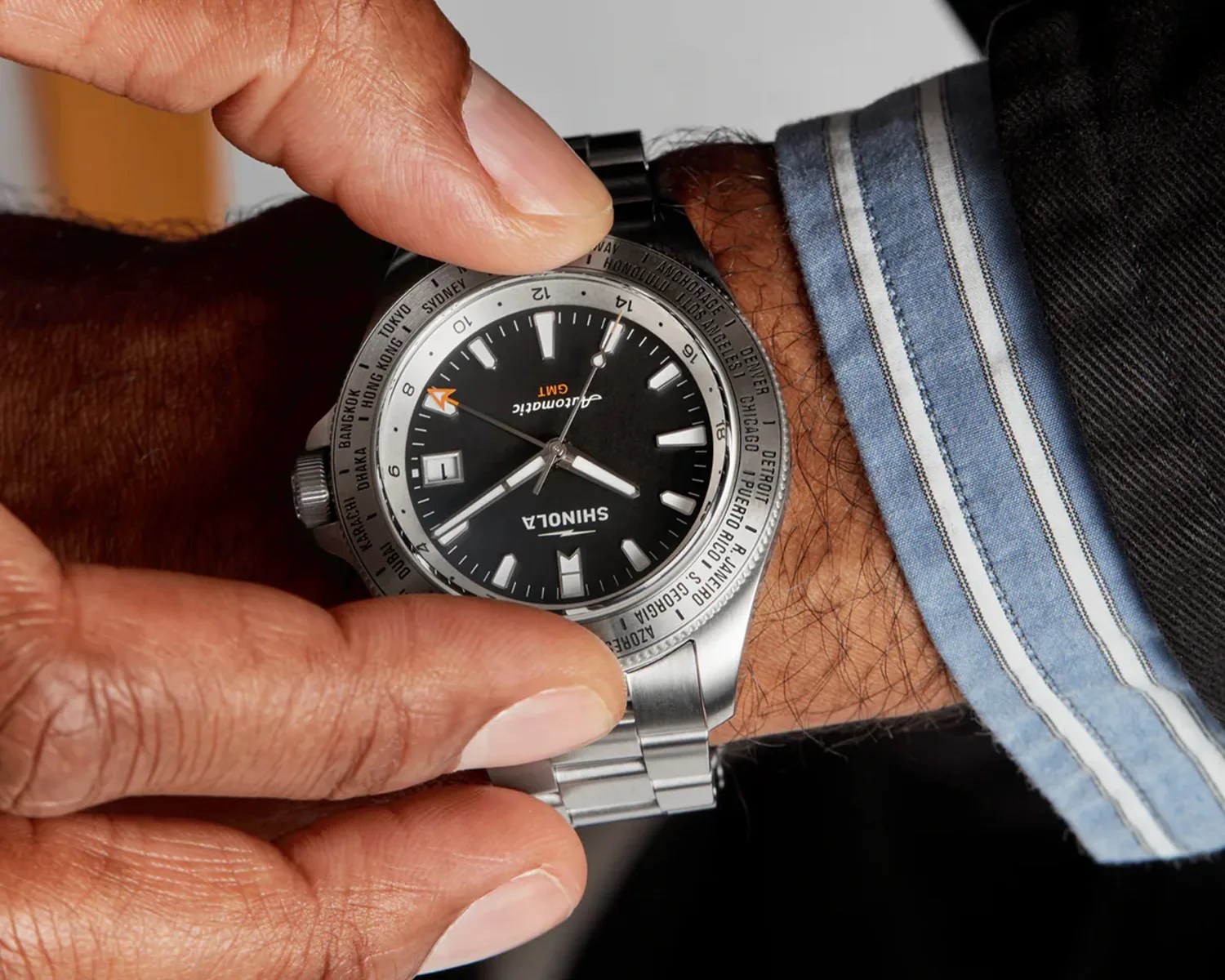 Silver Shinola GMT wristwatch with black dial and world time bezel worn on a wrist with a blue striped shirt cuff.