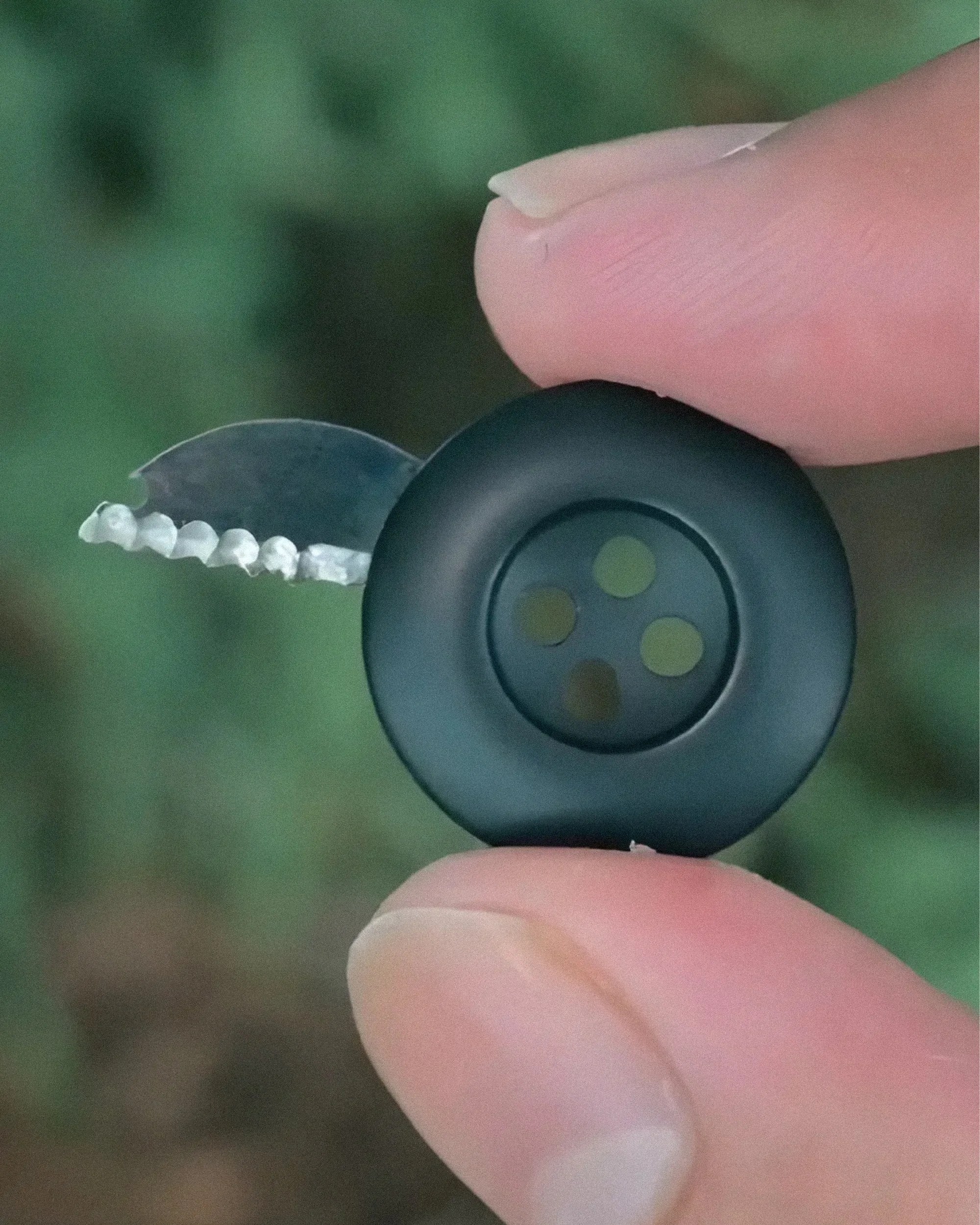 Black circular button with four holes held between fingers, with a small serrated metal blade partially extended.