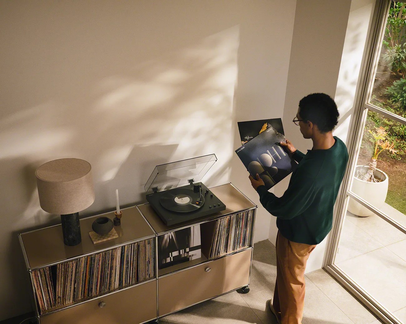 Person holding vinyl records next to a beige cabinet with a turntable and lamp near a window.