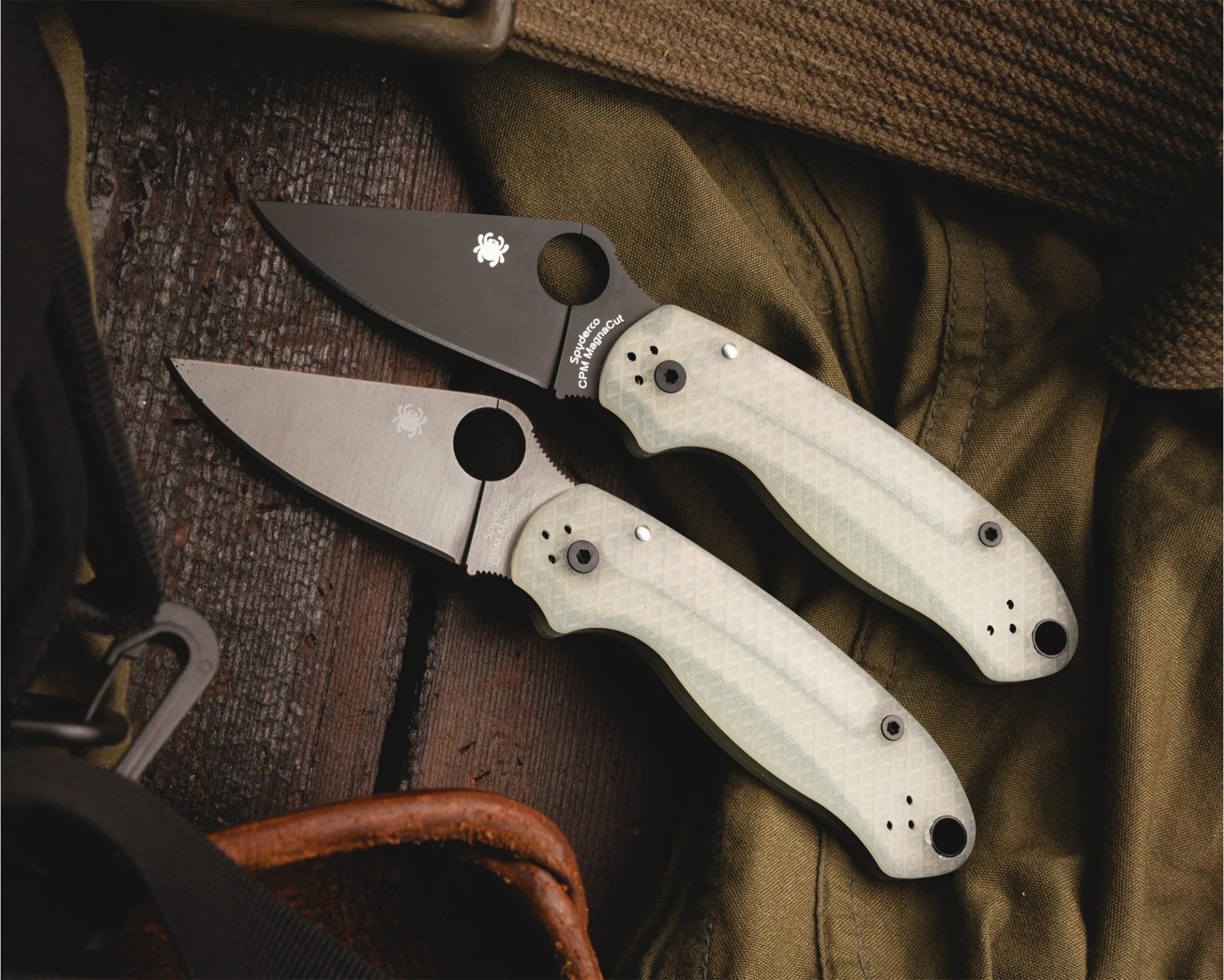 Two Spyderco folding knives with translucent white textured handles, one with a black blade and one with a silver blade, on a wooden surface and green fabric.