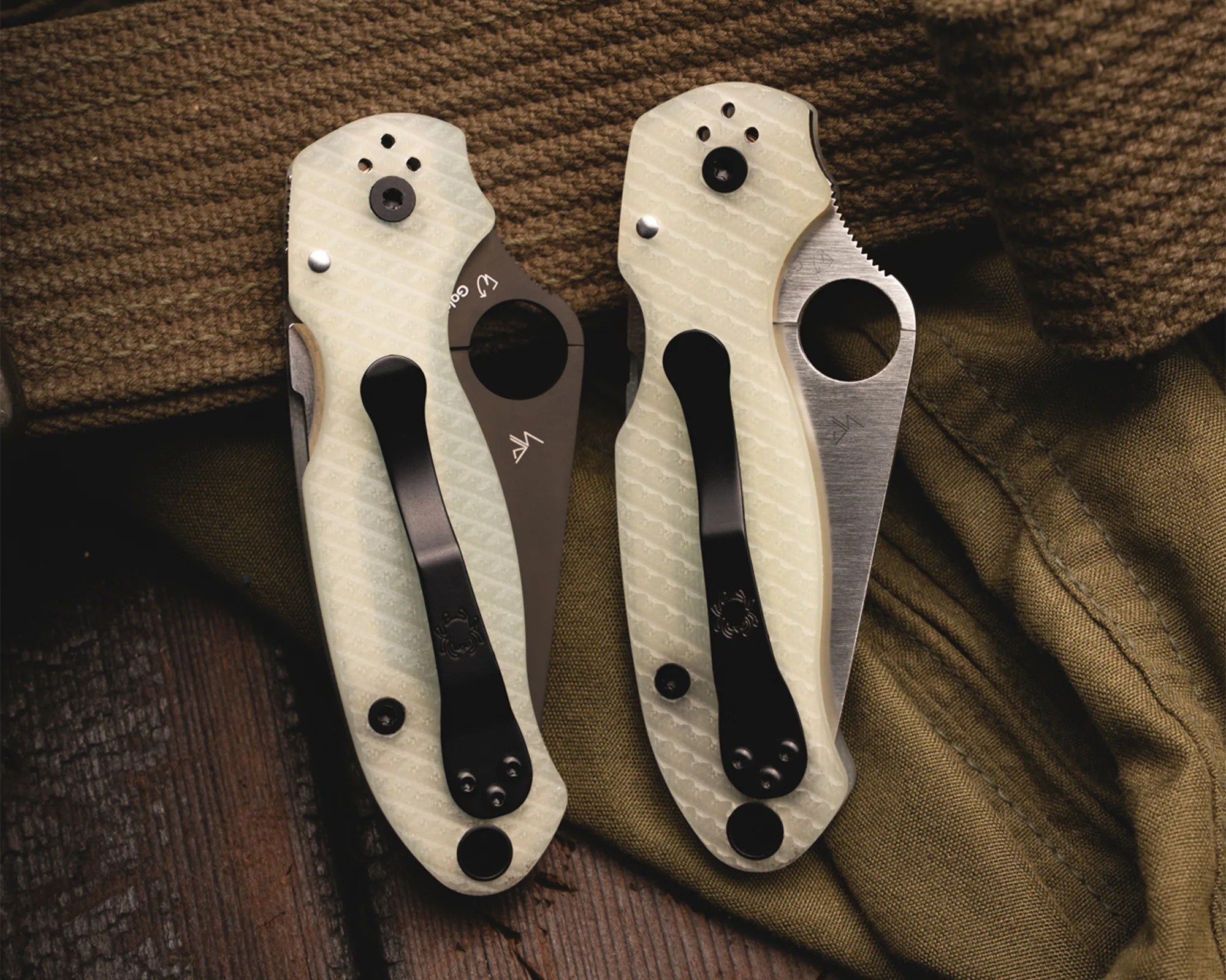 Two folding knives with textured white handles and black pocket clips resting on brown and olive fabric.