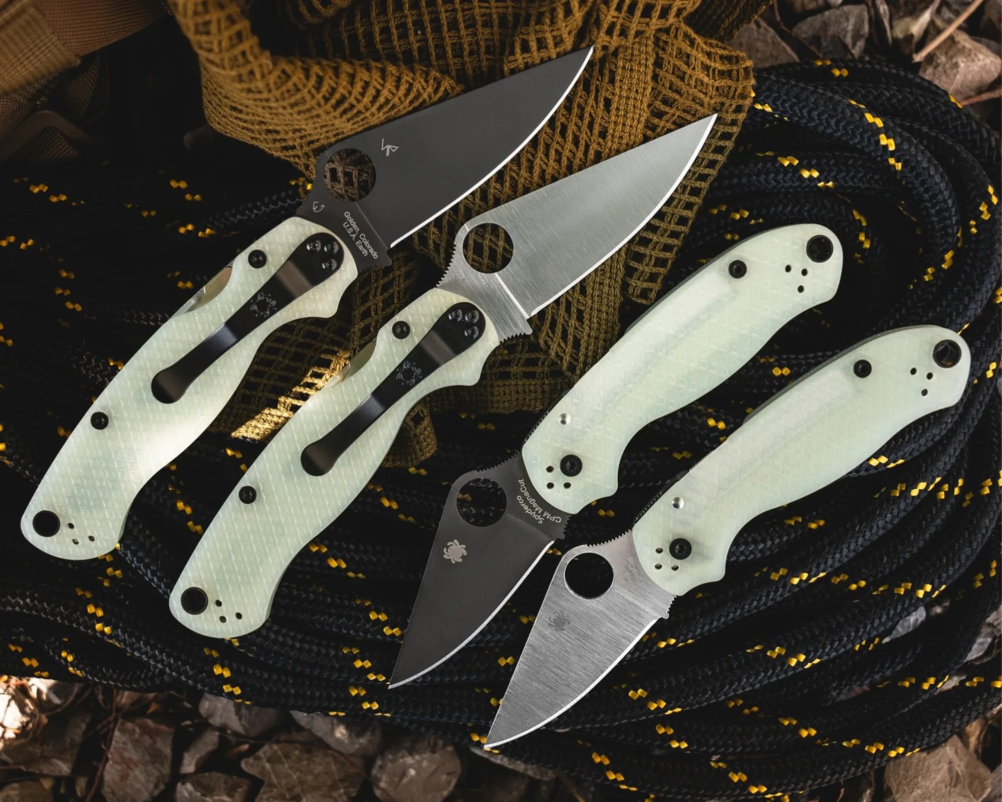 Four folding knives with light green textured handles and black or silver blades arranged on coiled black rope with yellow accents.