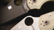 Close-up of two Spyderco CPM MagnaCut folding knives, one with a black blade and the other with a silver blade, both with light-colored handles.