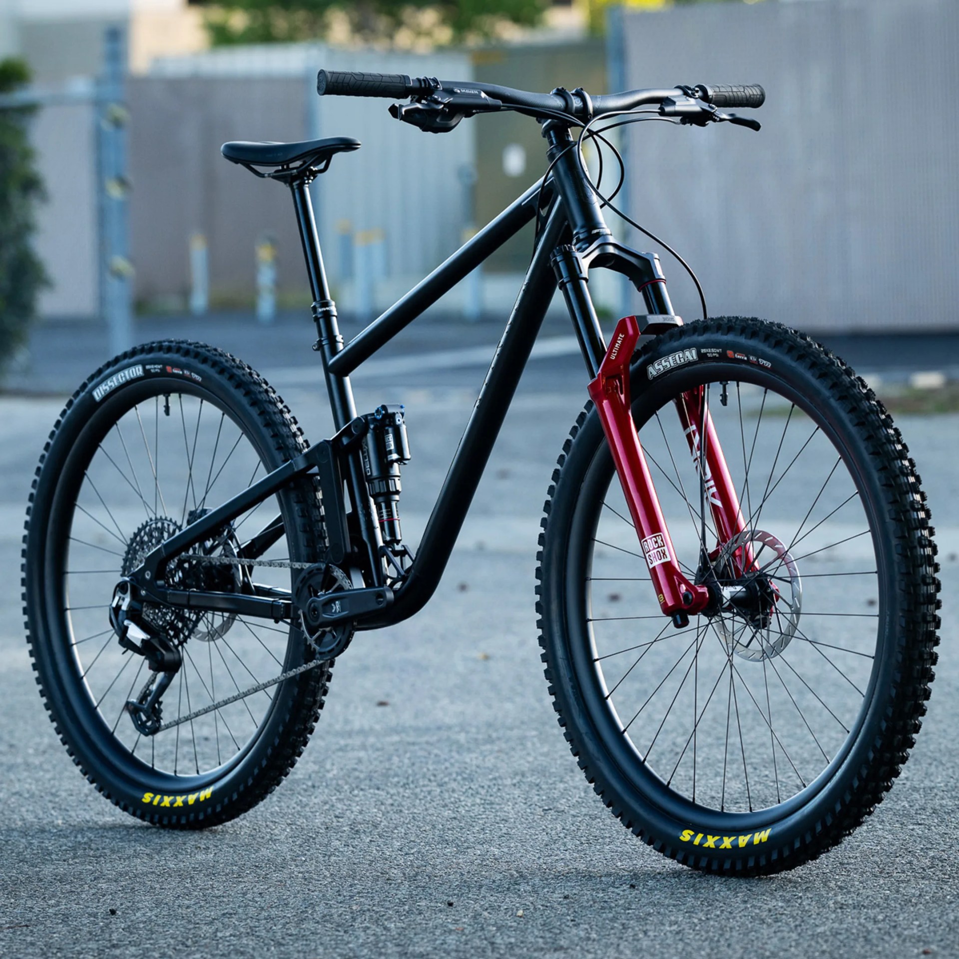 Matte black mountain bike with red RockShox front suspension fork and Maxxis tires on pavement.