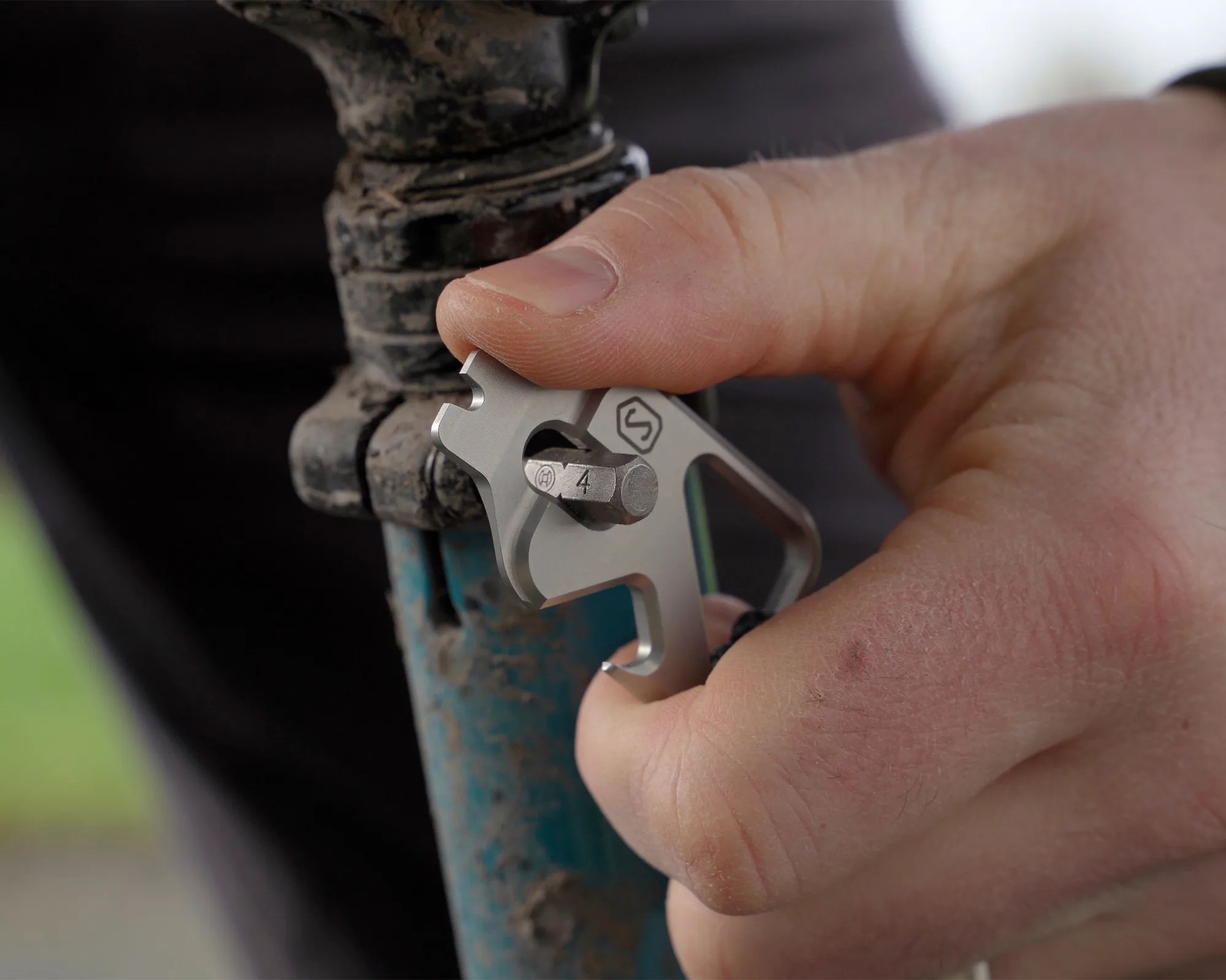 Hand holding a silver multi-tool tightening a bolt on a muddy blue bike frame.