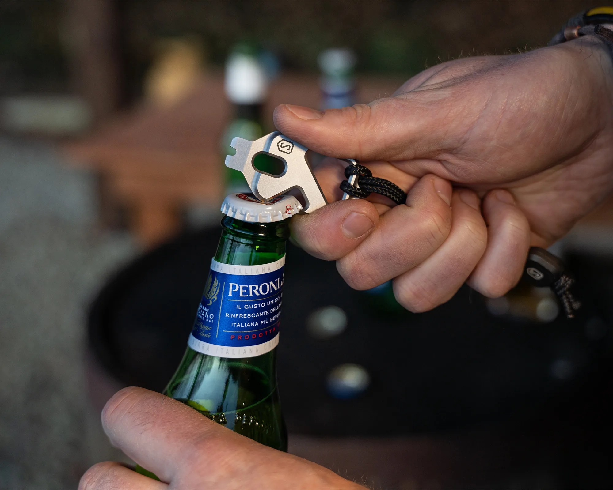 Hand using a silver multi-tool with a black lanyard to open a Peroni beer bottle.