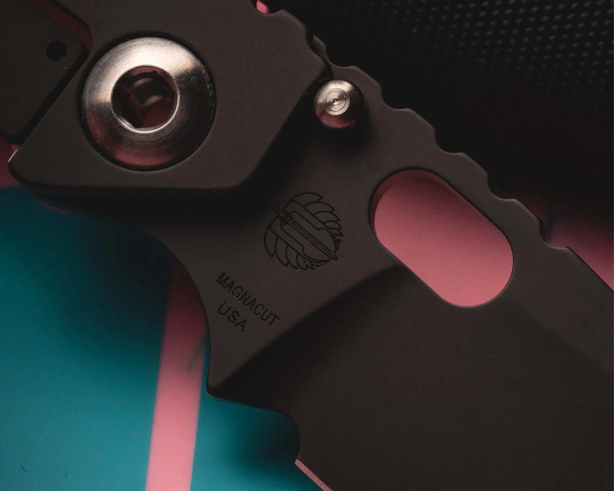 Close-up of a black metal tool or component with a circular silver part and engraved text "MAGNACUT USA" on a teal and pink background.