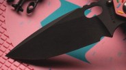Black tactical knife blade with serrated edge and "M. STRIDER" engraved on the handle, on a pink and blue surface.