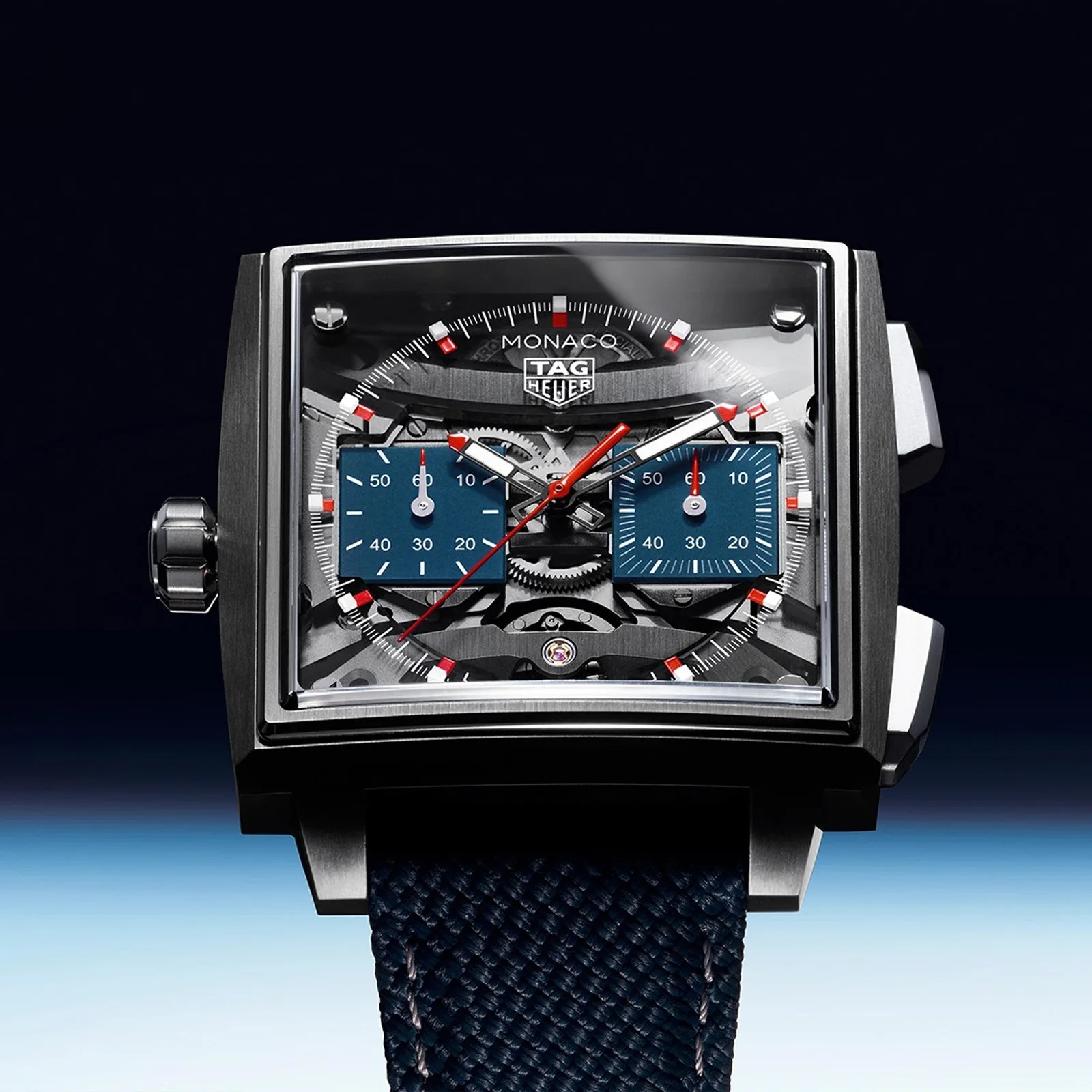 Square TAG Heuer Monaco watch with black case, transparent dial showing gears, blue subdials, red and white hands, and blue textured strap.