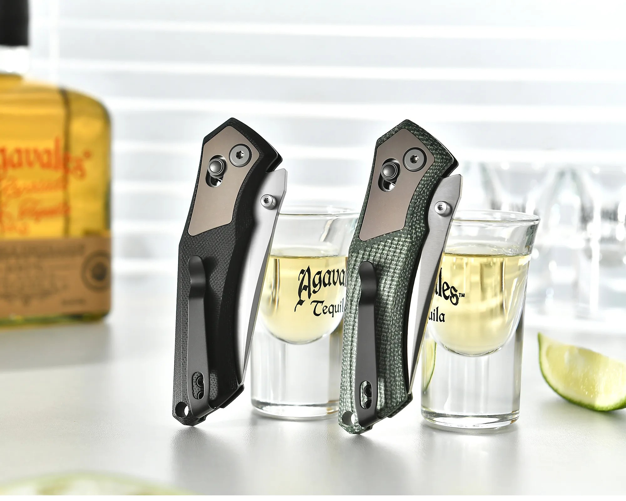 Two folding pocket knives, one black and one green, standing upright between two tequila shot glasses with lime wedge on white surface.