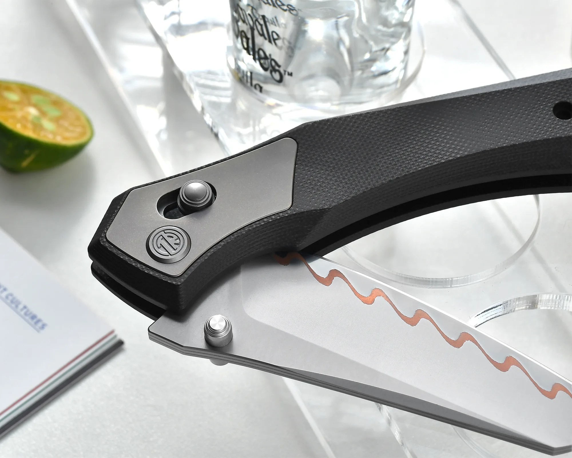 Close-up of a folding knife with a black textured handle and a silver blade featuring a wavy copper-colored pattern.