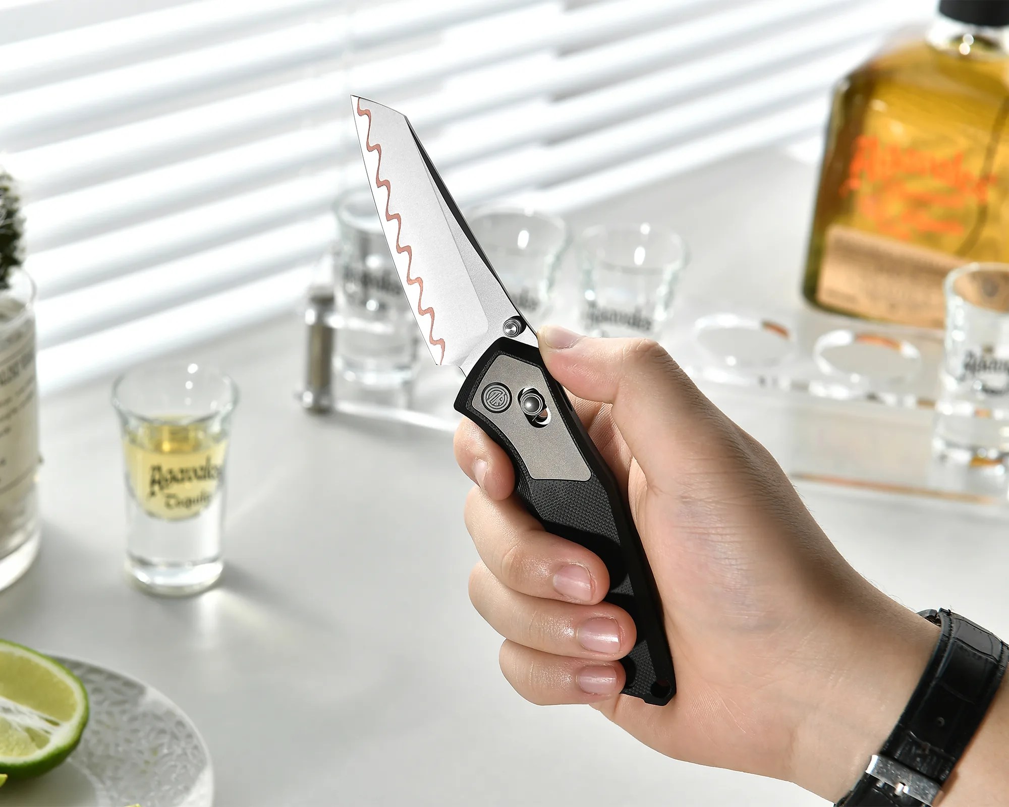Hand holding a folding knife with a wavy-edged blade and black textured handle over a white table with shot glasses and lime wedges.