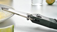 Close-up of a folding knife with a textured green handle and partially open blade resting near a plate and lime wedges.