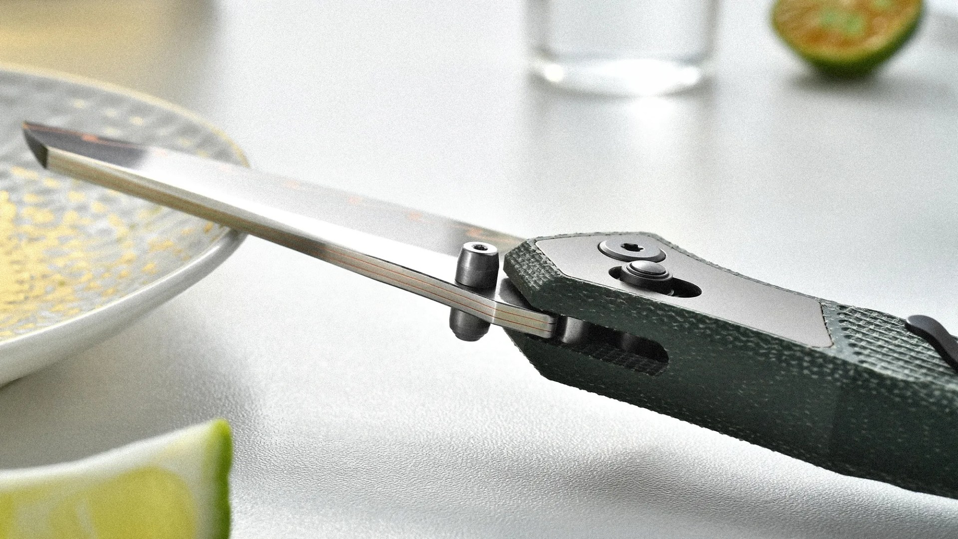 Close-up of a folding knife with a textured green handle and partially open blade resting near a plate and lime wedges.