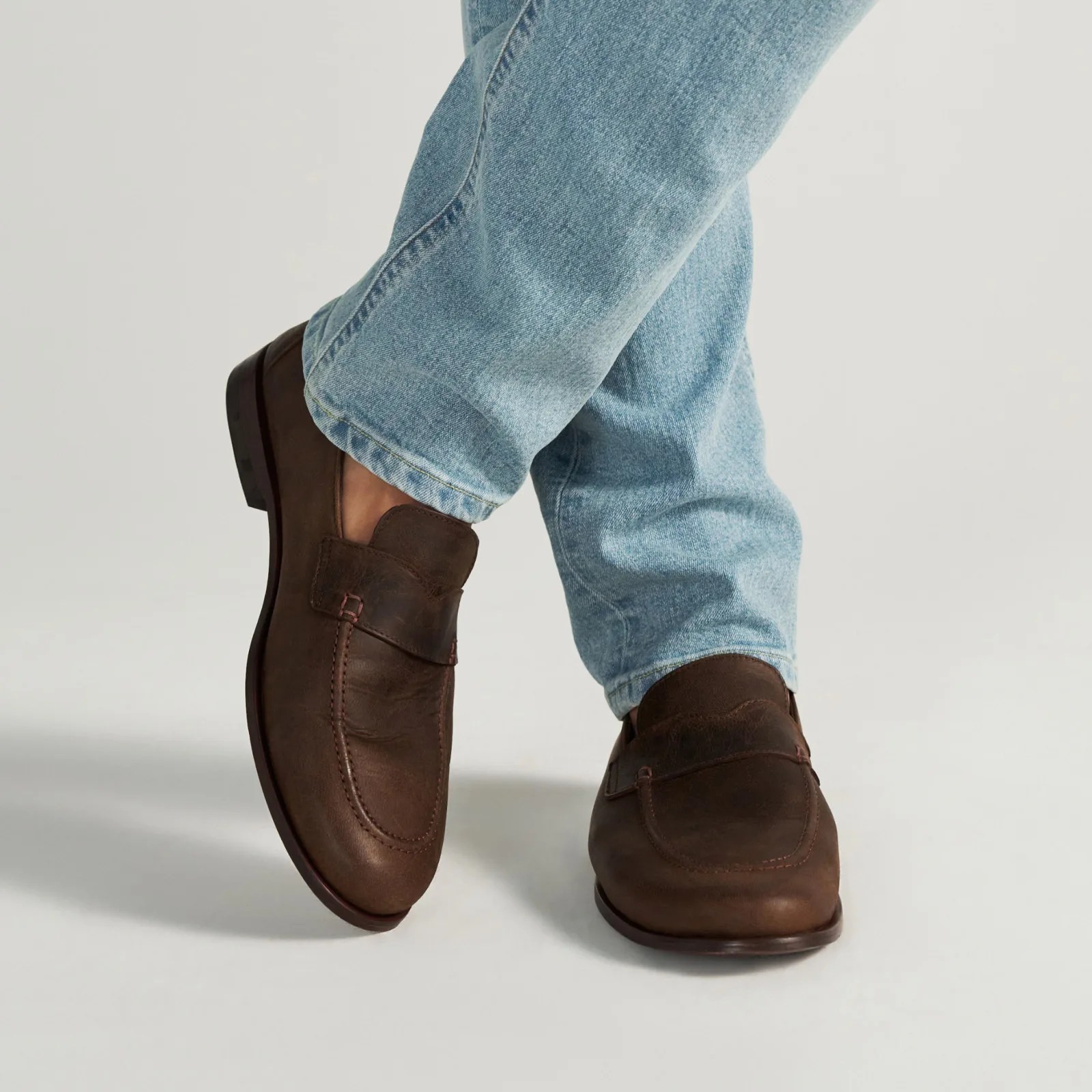 Brown leather loafers worn with light blue denim jeans against a plain background.
