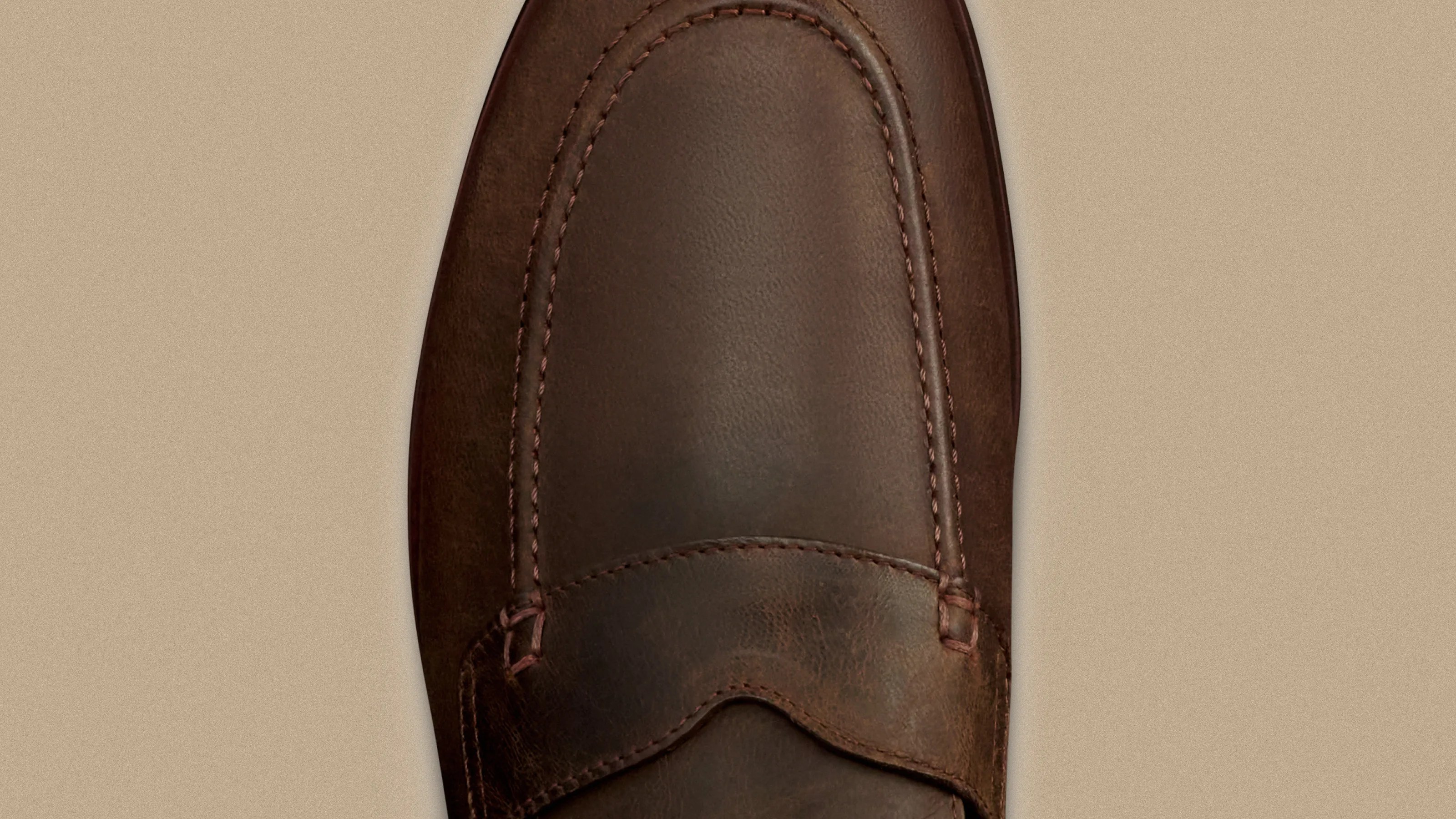 Close-up of the toe area of a brown leather loafer with detailed stitching on a beige background.
