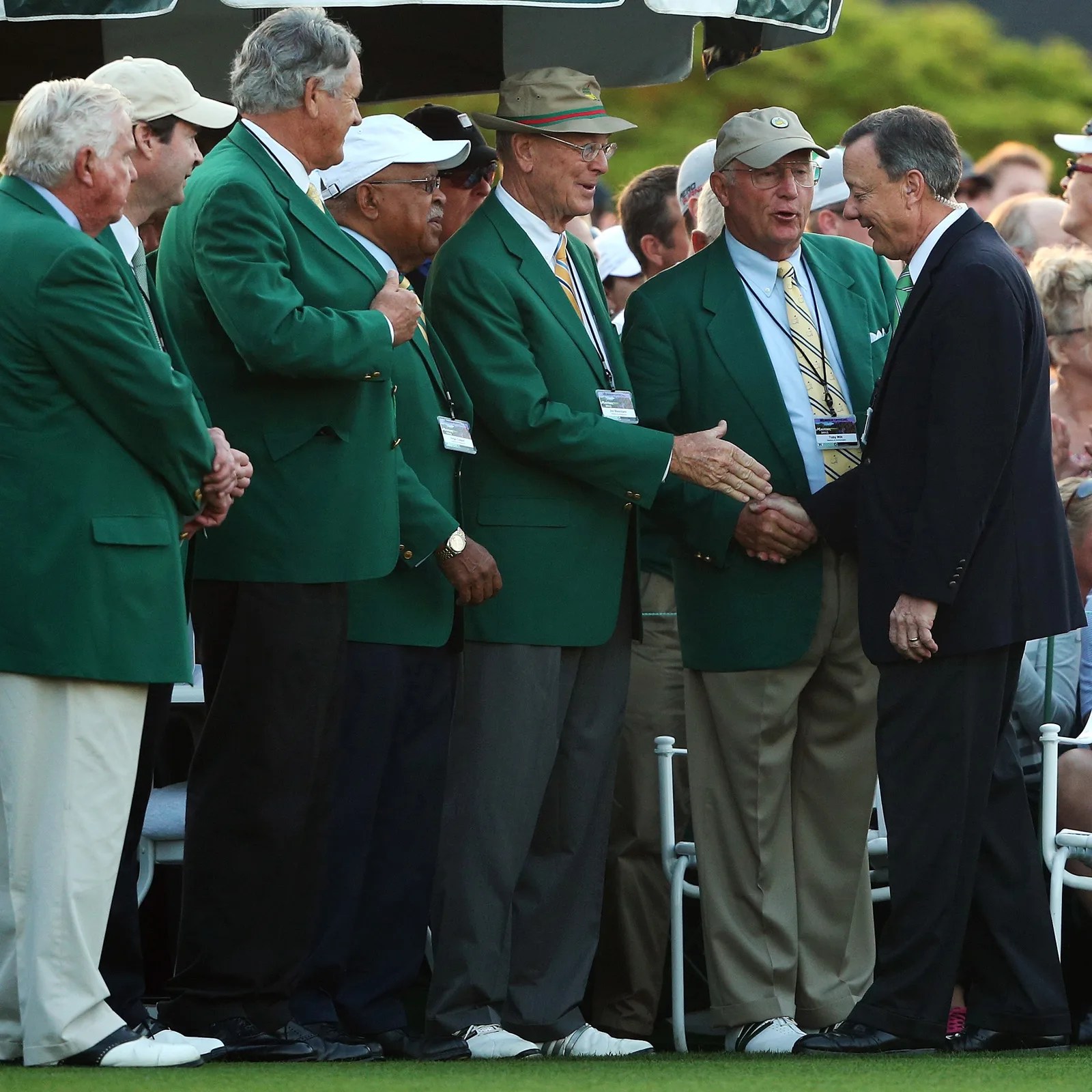 Men in green jackets and dress pants shaking hands with a man in a dark suit on a golf course.