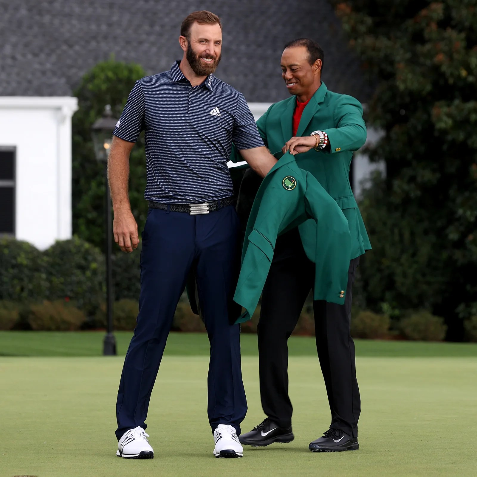 Golfer in navy pants and striped Adidas polo receiving a green jacket from another man in a green jacket on a golf course.