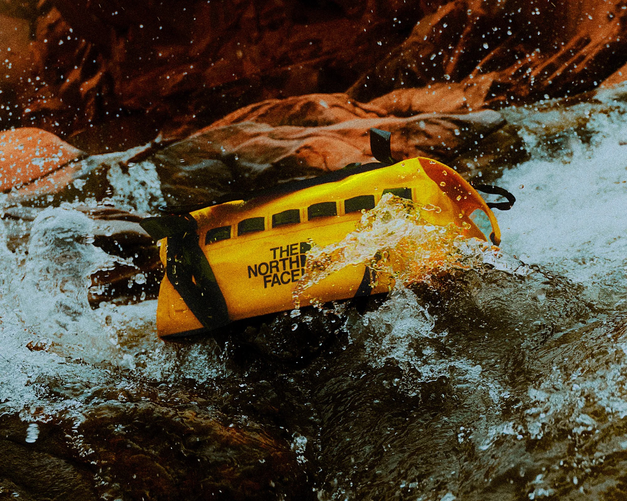 Yellow waterproof The North Face dry bag floating in splashing river water near rocks.