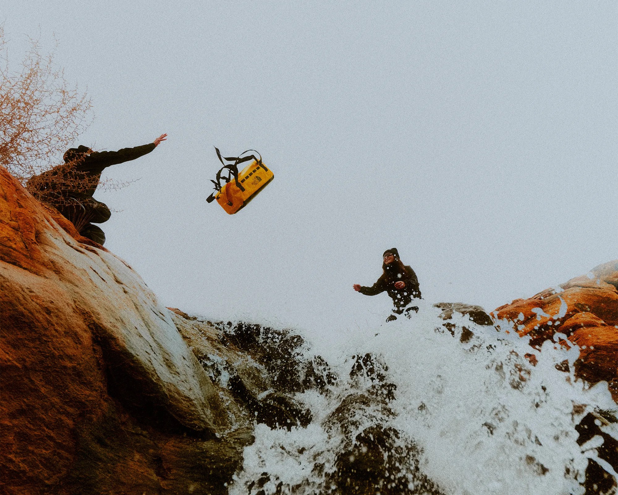 Yellow duffel bag being tossed between two people on rocky cliffs above a waterfall.
