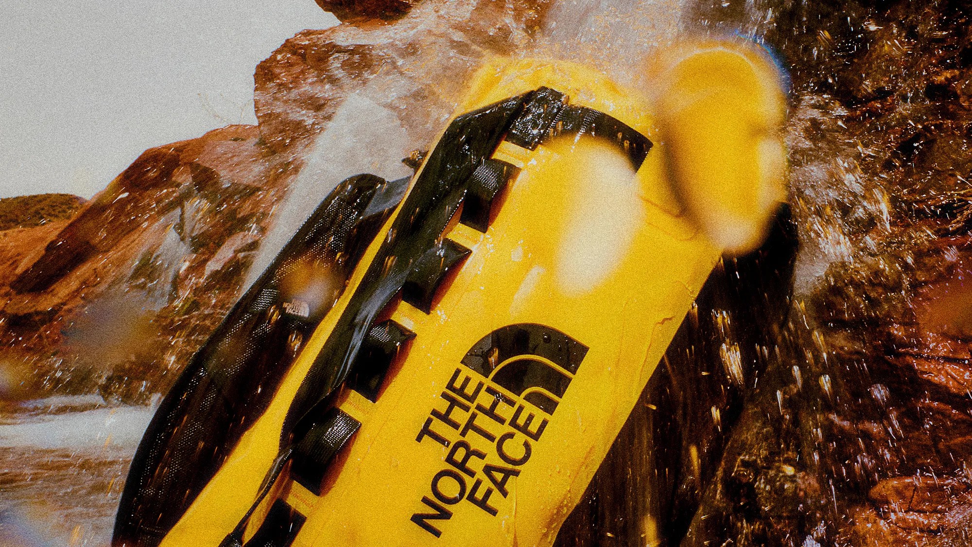 Yellow waterproof dry bag with black straps and The North Face logo, splashed with water near rocks.