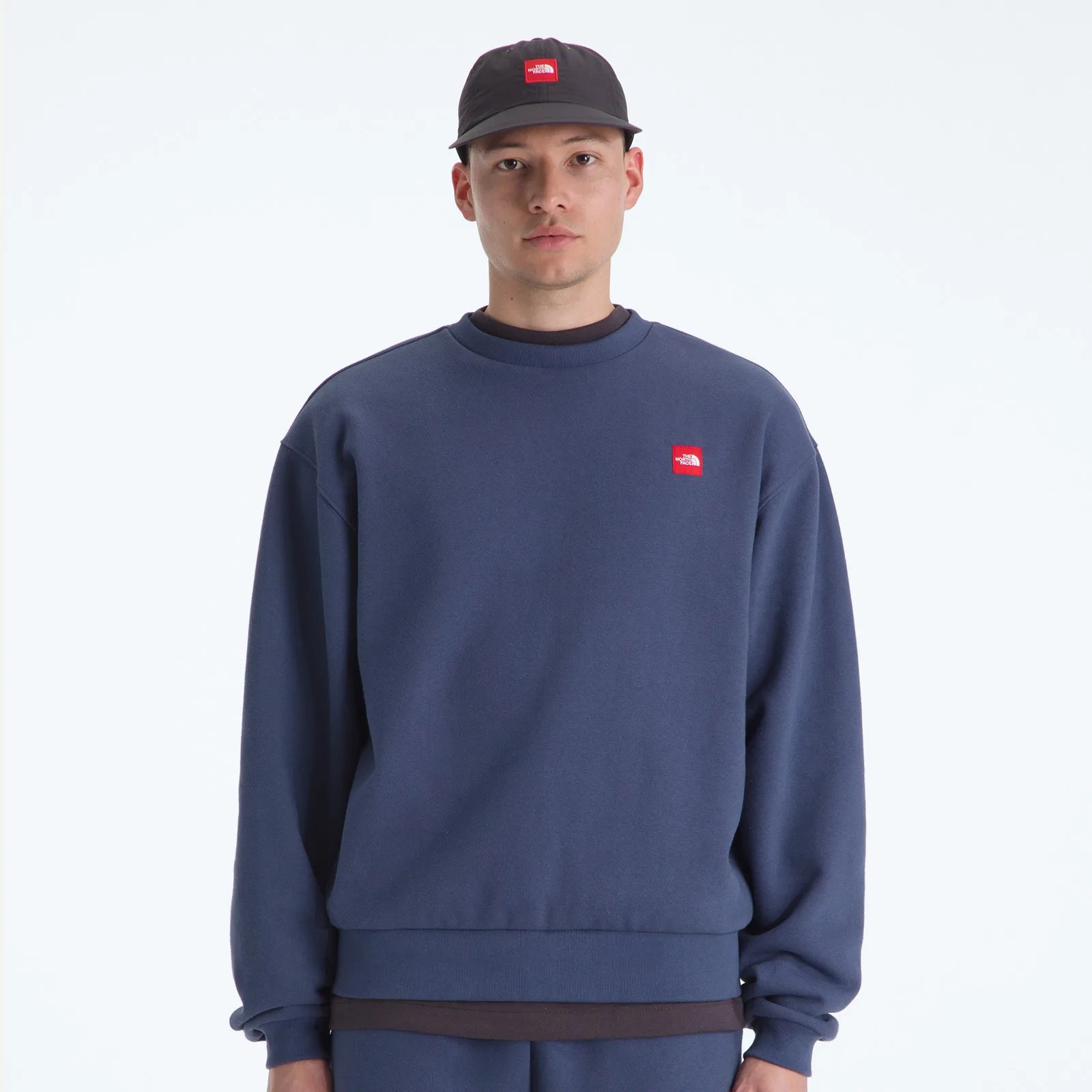 Man wearing a navy blue crewneck sweatshirt and matching cap with red square logos on the chest and front.