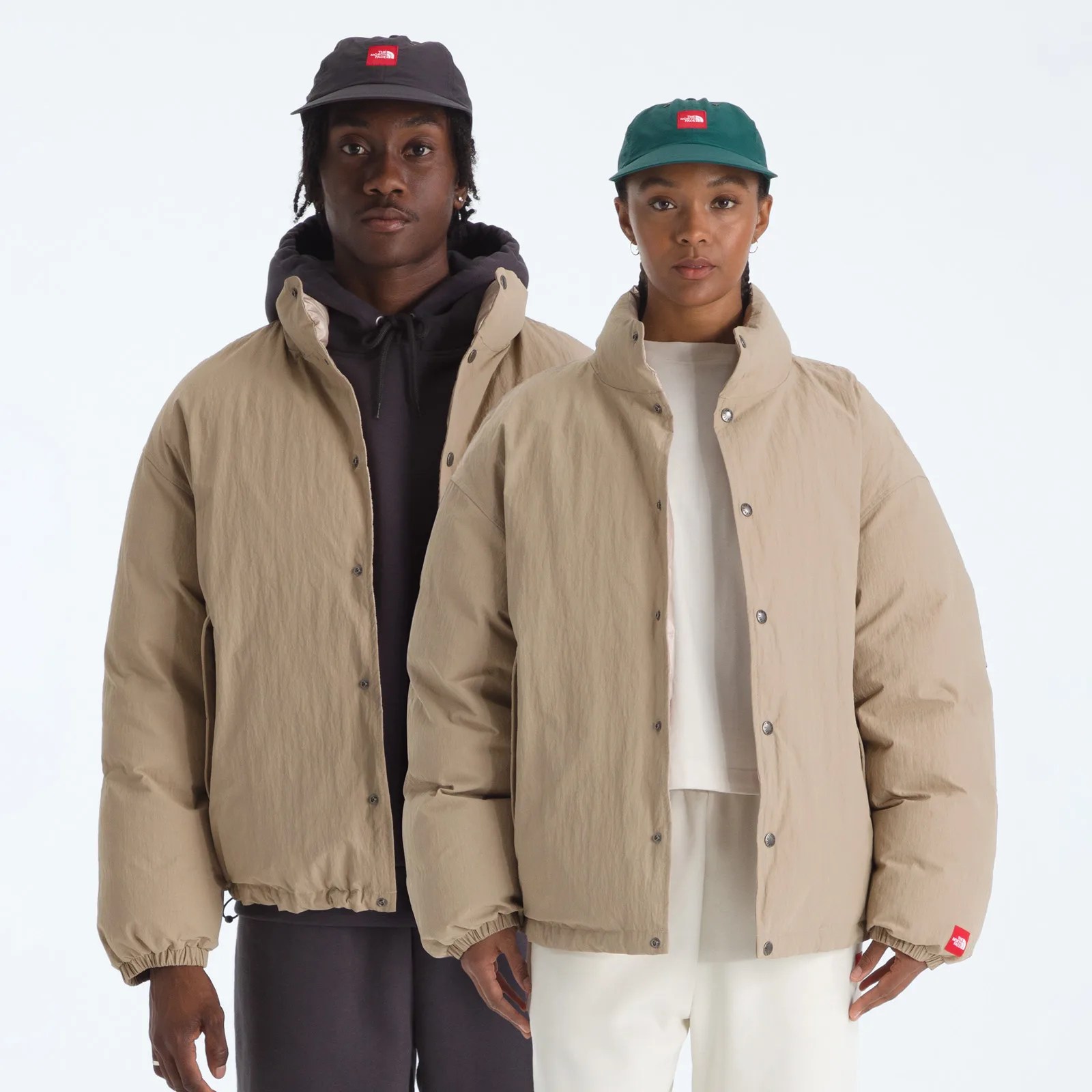 Two people wearing beige puffer jackets and The North Face caps, one black and one green, standing against a white background.