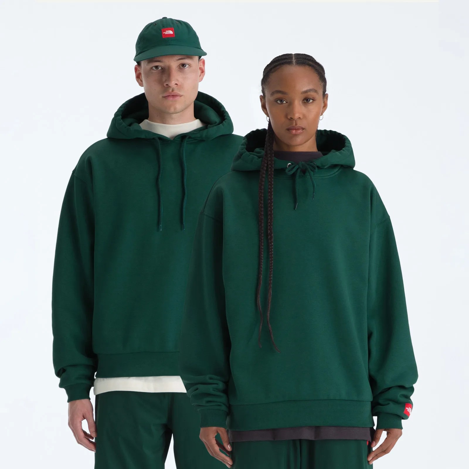 Two people wearing matching dark green hoodies and pants, one also wearing a dark green cap with a red logo patch.