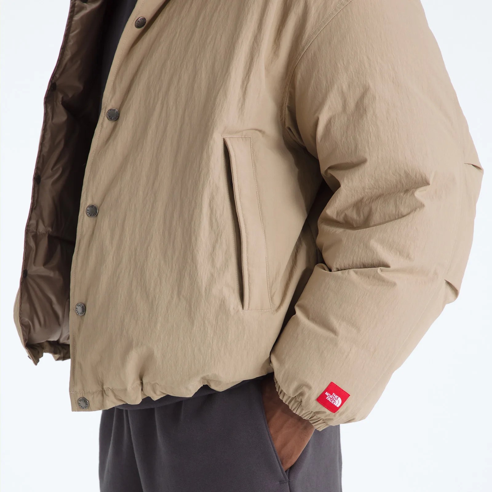 Beige puffer jacket with snap buttons and a red The North Face logo patch on the cuff, worn with dark pants.