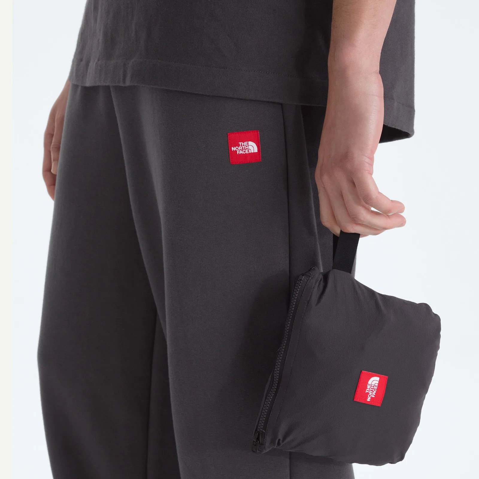 Close-up of a person wearing dark gray The North Face pants and holding a matching black pouch with a red logo patch.