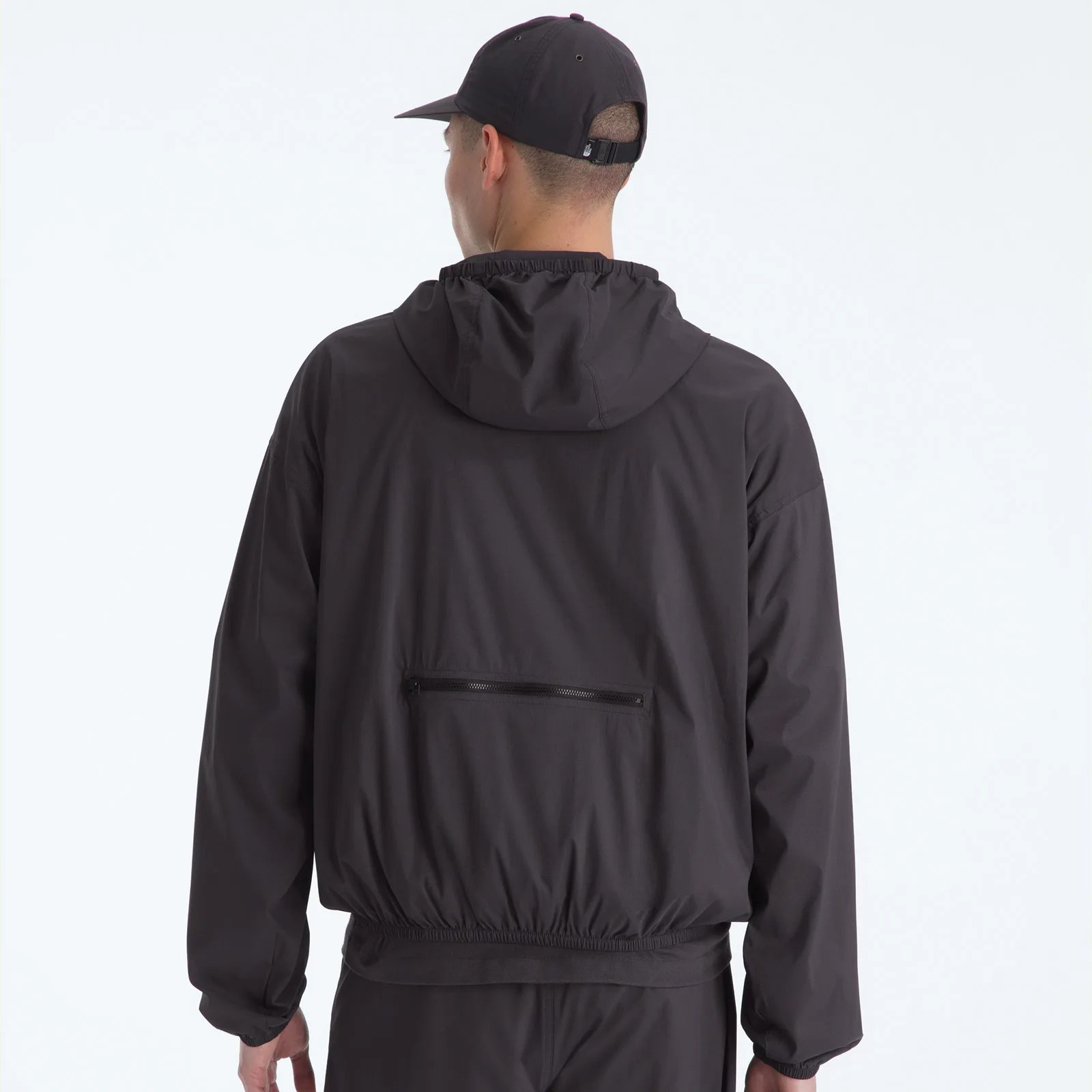 Man wearing a black hooded jacket with a horizontal zippered pocket on the back and a black cap.