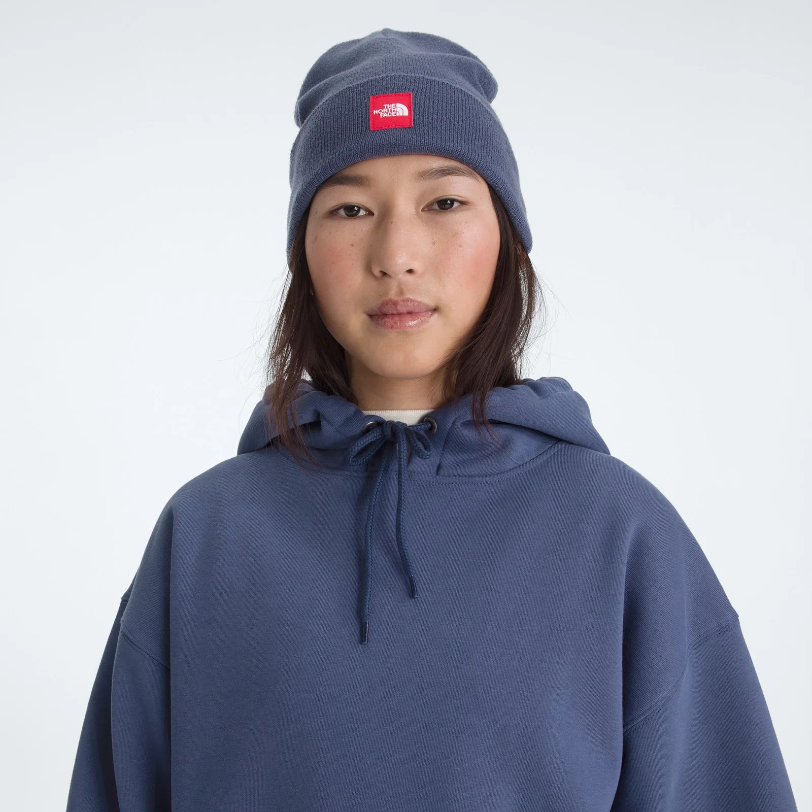 Person wearing a navy blue knit beanie with a red The North Face logo patch and a matching navy blue hoodie.