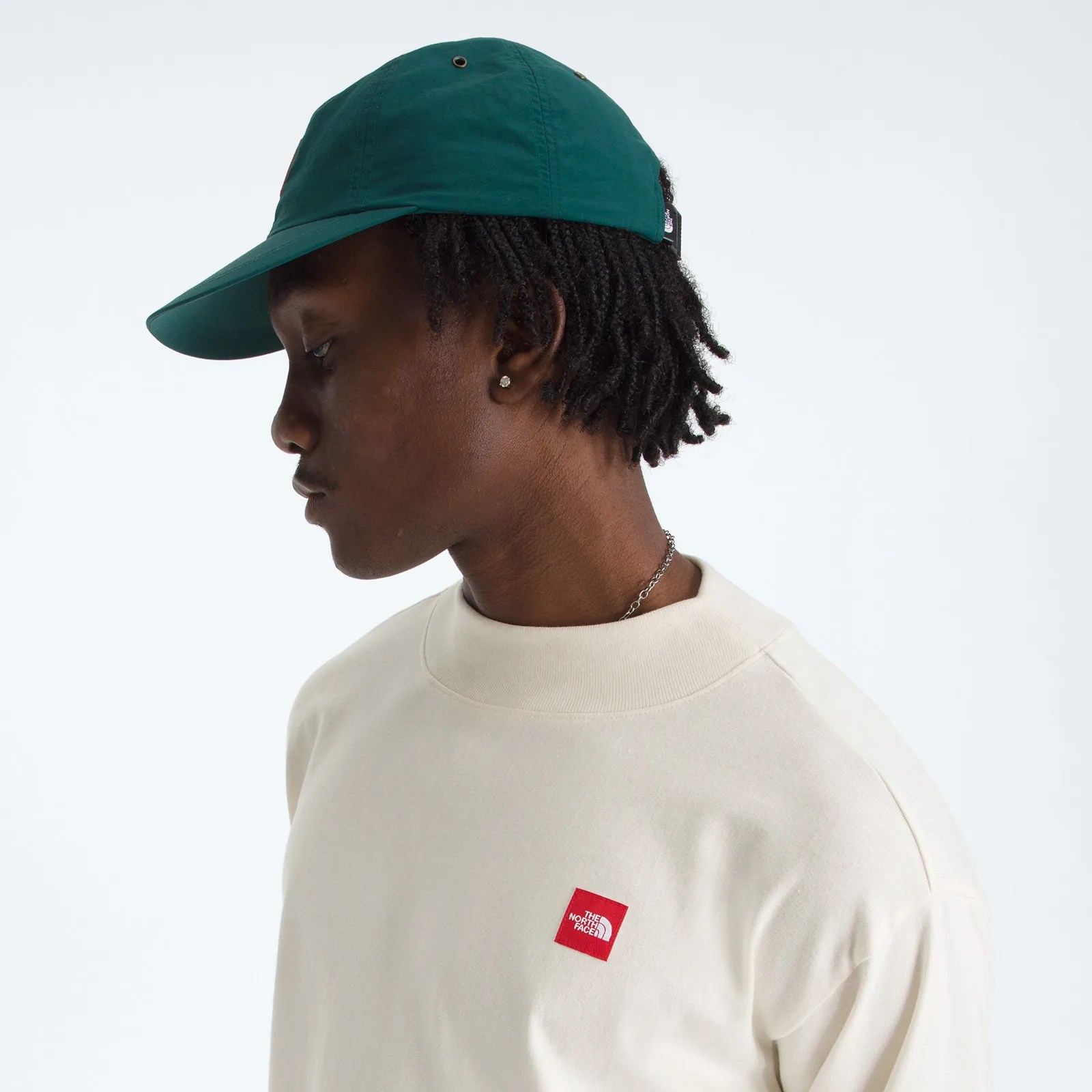 Side view of a person wearing a dark green cap and an off-white sweatshirt with a small red The North Face logo on the chest.