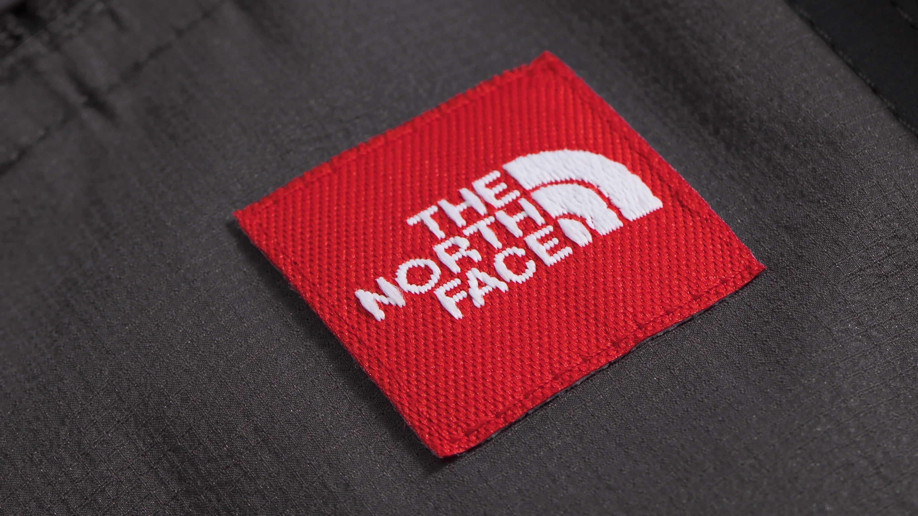 Red square fabric patch with white "The North Face" logo sewn onto black textured fabric.