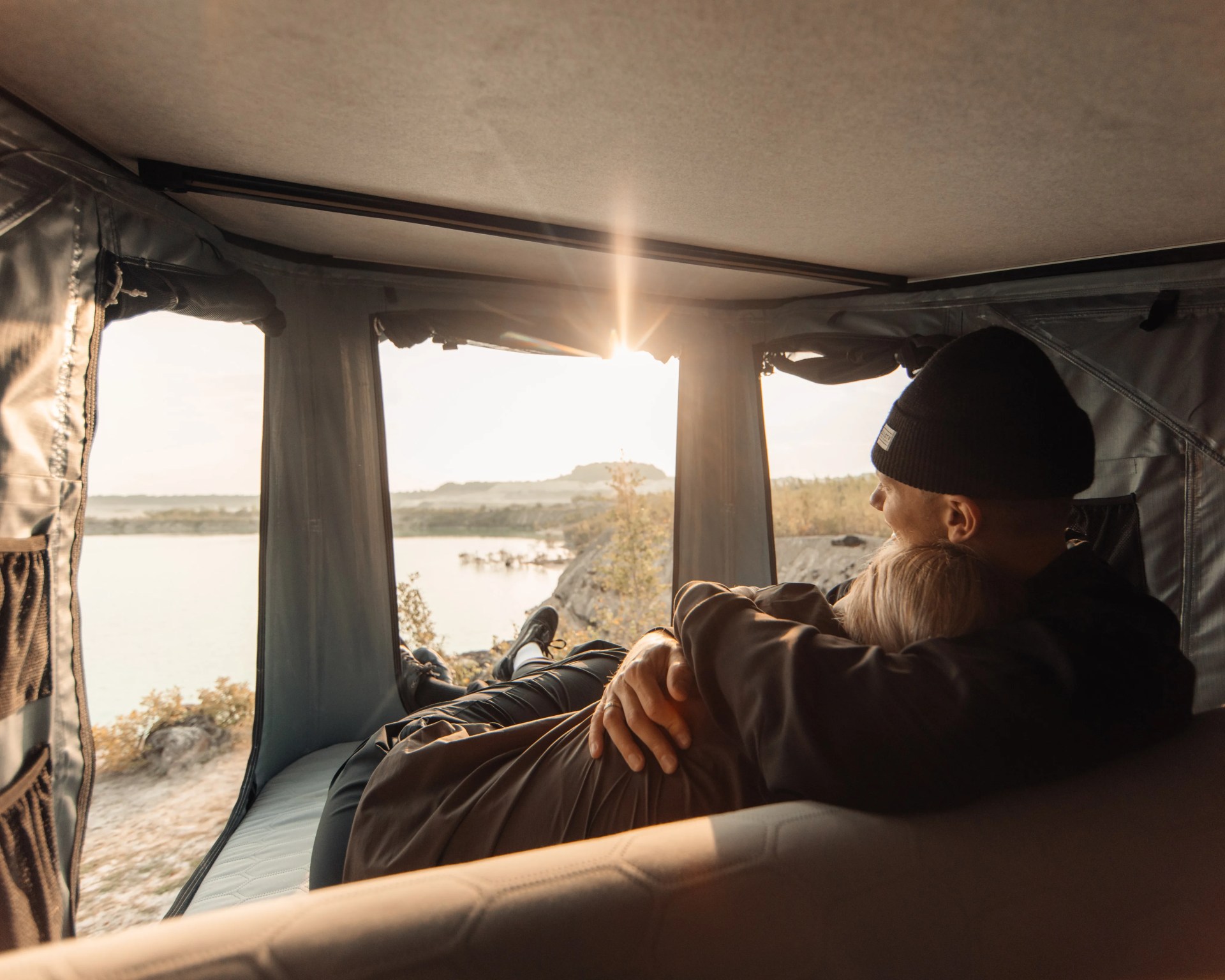 Two people embracing inside a tent with a view of a lake and sunset outside.