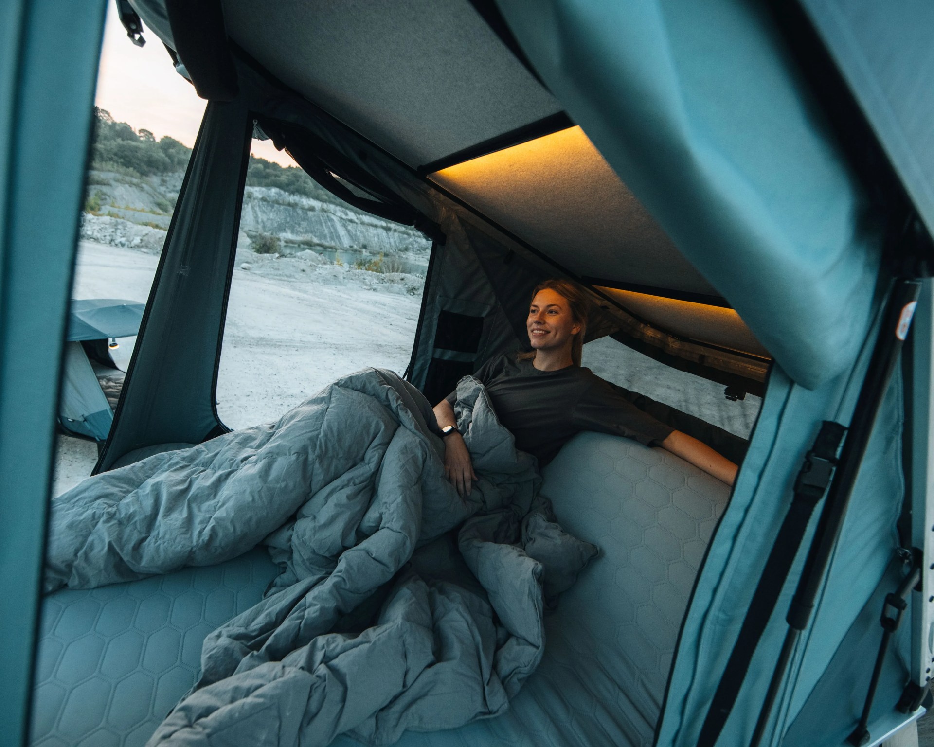 Person lying inside a rooftop tent with a gray sleeping bag and mattress, with outdoor rocky terrain visible outside.
